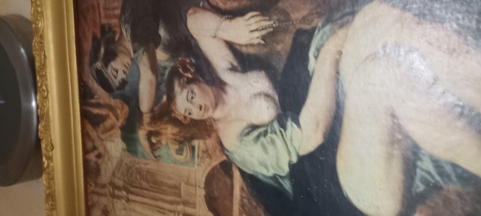 The image is a close-up of a framed painting. The painting depicts a scene with at least two individuals. In the foreground, a woman is seated, facing the viewer, with her torso exposed. She has fair skin, light brown hair styled with a flower, and is wearing a green garment draped over her shoulder. Her right arm is extended, and her hand is visible. Behind her, another person, possibly a man, with darker hair and a headband, is reaching over and appears to be attending to her hair. The background of the painting shows architectural elements that resemble classical ruins, with columns and arches. The overall style suggests it is a classical or Baroque painting. The frame is ornate and gold-colored. The image is captured at an angle, and the lighting suggests it is an indoor setting. There is no information to suggest this image is located in Kryvyi Rih, Ukraine.