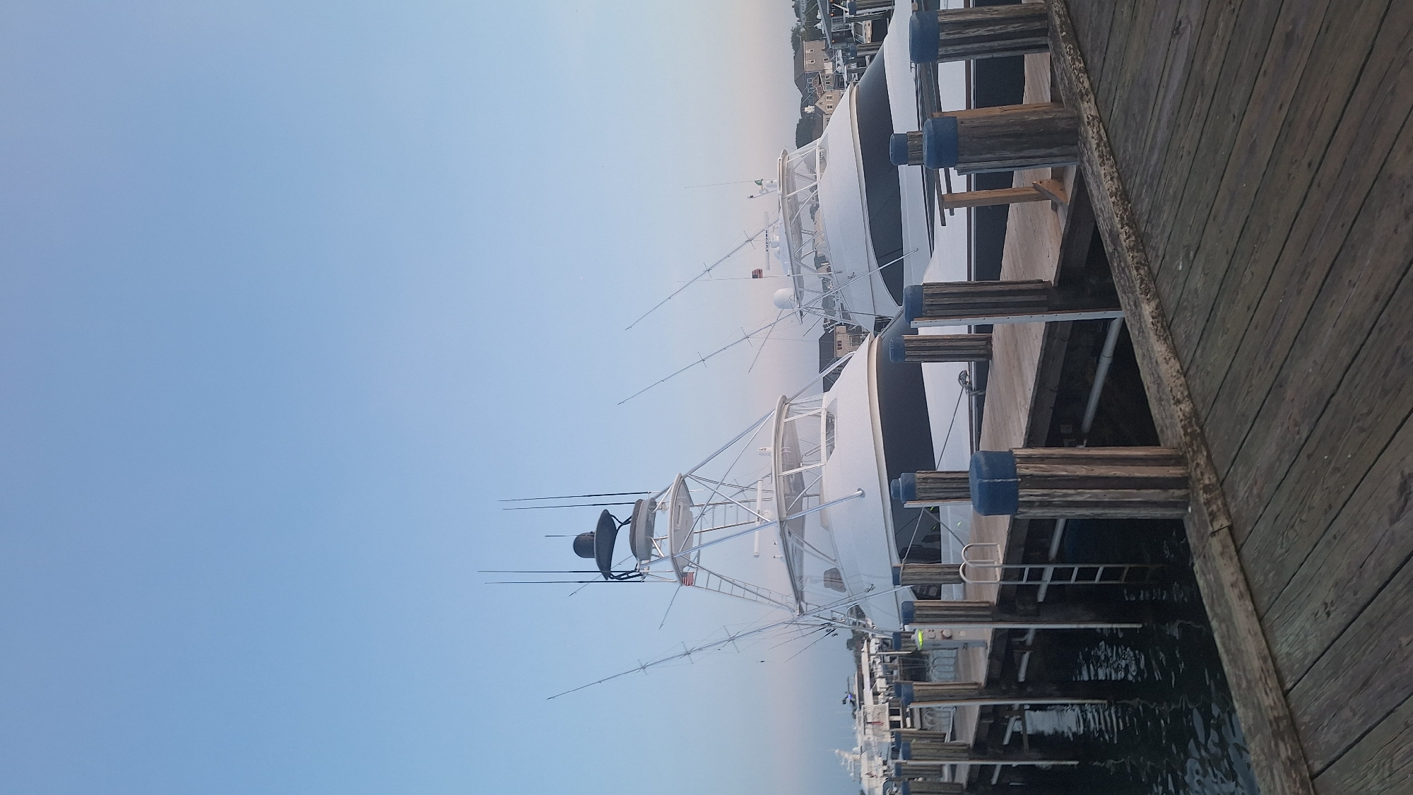 This is a medium-angle, vertical, outdoor, eye-level photograph of two white yachts docked side-by-side at a wooden pier. The sky is a clear, pale blue with no clouds. The yachts are large, white vessels with dark trim along the hull. Both have tall, silver towers with multiple outriggers, suggesting they are fishing boats. The boat on the left is closer to the camera and appears to be slightly larger. The pier is made of weathered wooden planks and is supported by thick, wooden pilings topped with blue caps. The water around the pier is dark and still. In the background, beyond the yachts, there is a cluster of houses and more boats docked. The scene appears to be at dusk or dawn. The image was taken in Nantucket, United States of America.