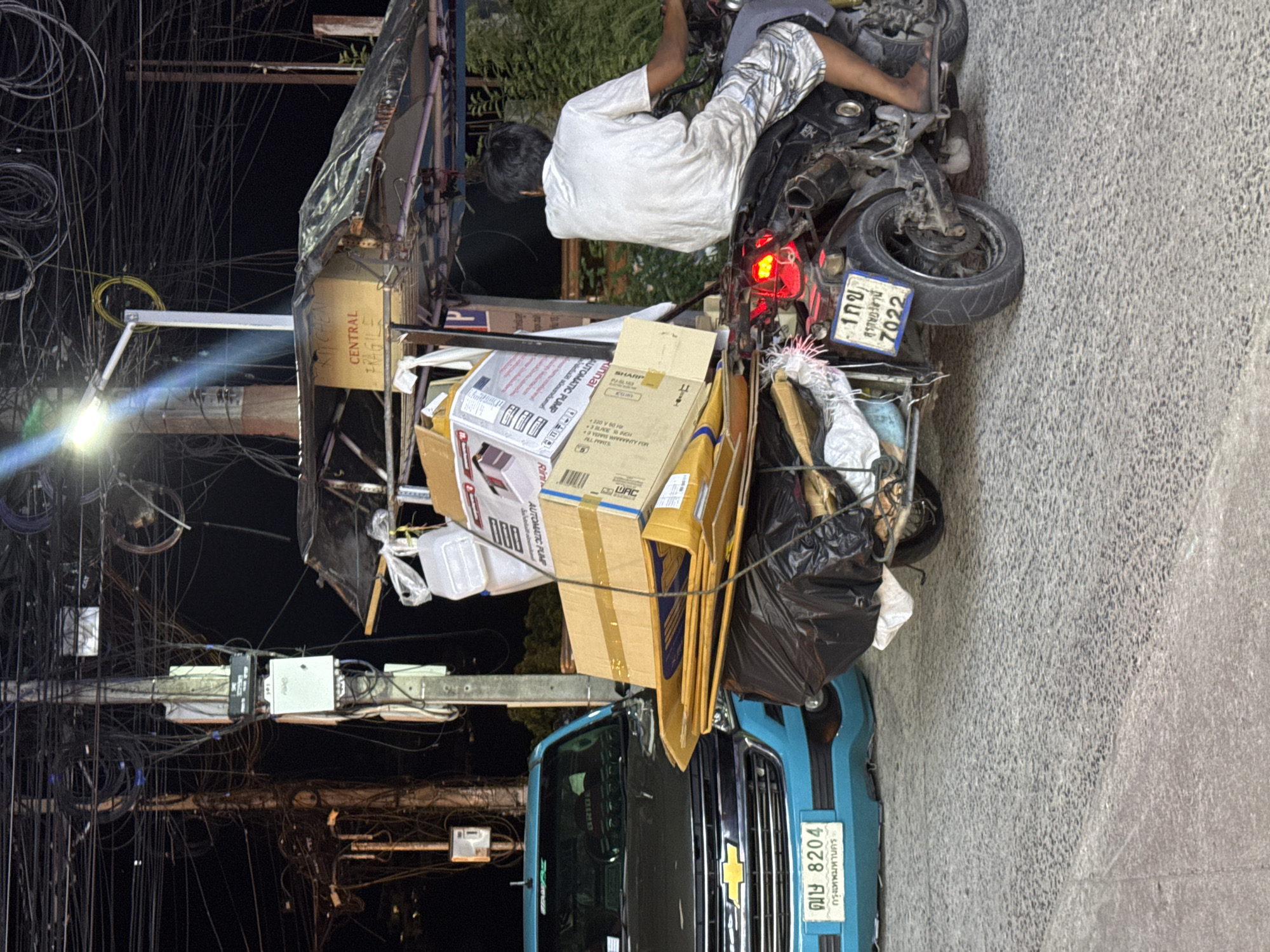 The image depicts a nighttime scene on a street, likely in Thailand, given the license plate on the blue vehicle and the Thai script. The primary subjects are a person sitting on a heavily loaded motorcycle, and a blue pickup truck parked nearby. The motorcycle is piled high with cardboard boxes and what appears to be refuse in black trash bags, suggesting it's being used for transport or disposal. The person is wearing a white shirt and patterned shorts.
The setting is characterized by a dense network of overhead electrical wires, common in urban Asian environments. A street light casts a bright beam across some of the wires. The ground is paved asphalt.
The presence of the loaded motorcycle and the truck, combined with the dark sky and street lighting, points to an activity related to delivery, moving, or waste collection happening at night. There are no overt emotional cues visible.
Visible text includes Thai characters on the license plates, reading "ฌษ 8204 กรุงเทพมหานคร" (Chor Sor 8204, Bangkok) on the truck and "1กข 7022" (1 Kor Khor 7022) on the motorcycle. Labels on the boxes are also visible, such as "KITCHEN CENTRAL FRAGILE," "AUTOMATIC PUMP," and "SHARP," indicating the contents are household items or appliances. The overall impression is one of bustling urban activity at night, with an emphasis on the practicalities of transport and logistics.