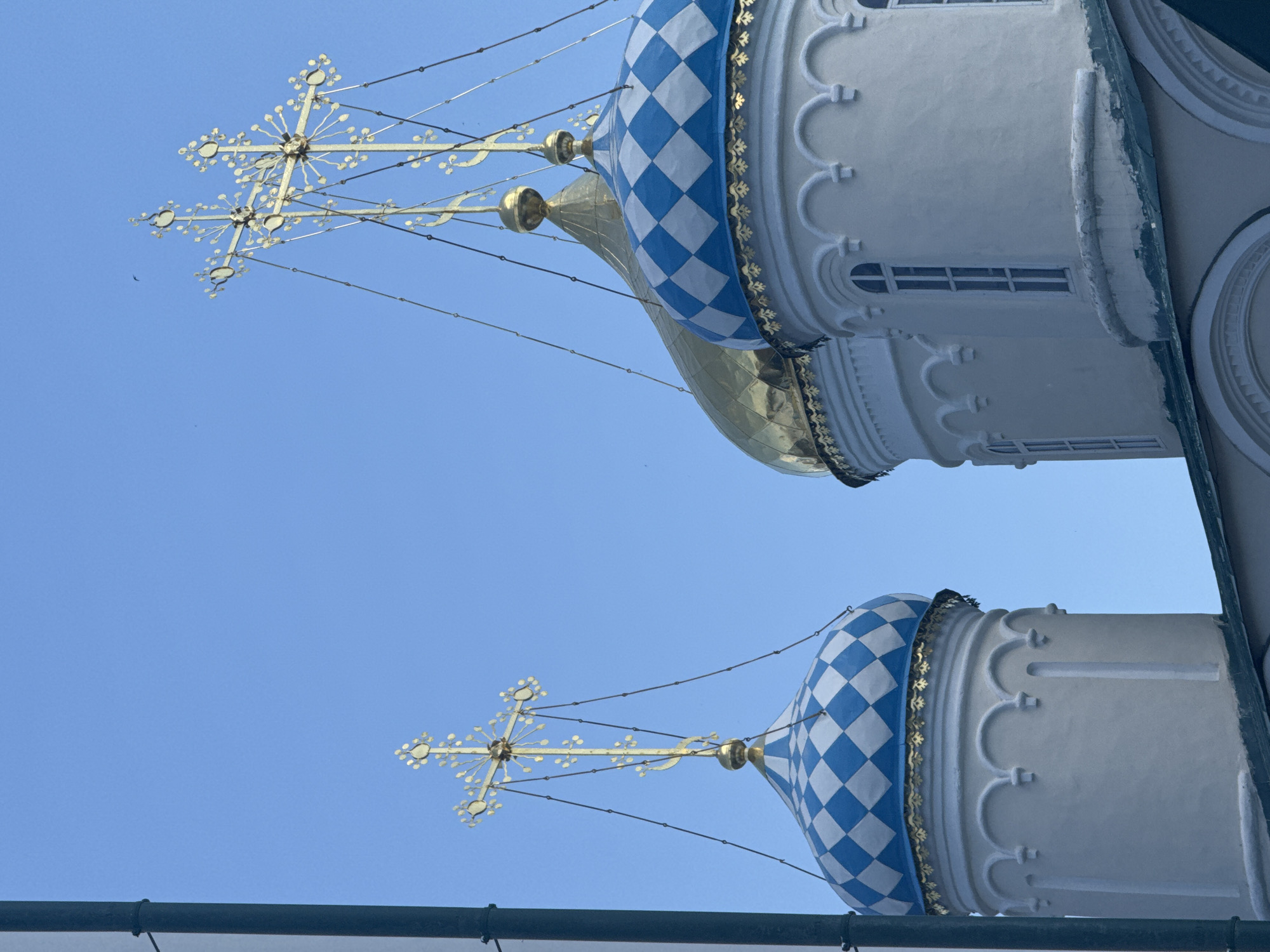 The image shows two onion domes of a Russian Orthodox church in Kazan, Russia, under a clear blue sky. The domes have a checkered pattern of blue and white tiles around the upper portion. Each dome is topped with a golden cross. The domes are part of a white building with architectural details such as rounded arches and small windows. The photograph is taken from a low angle, looking up at the domes and crosses.