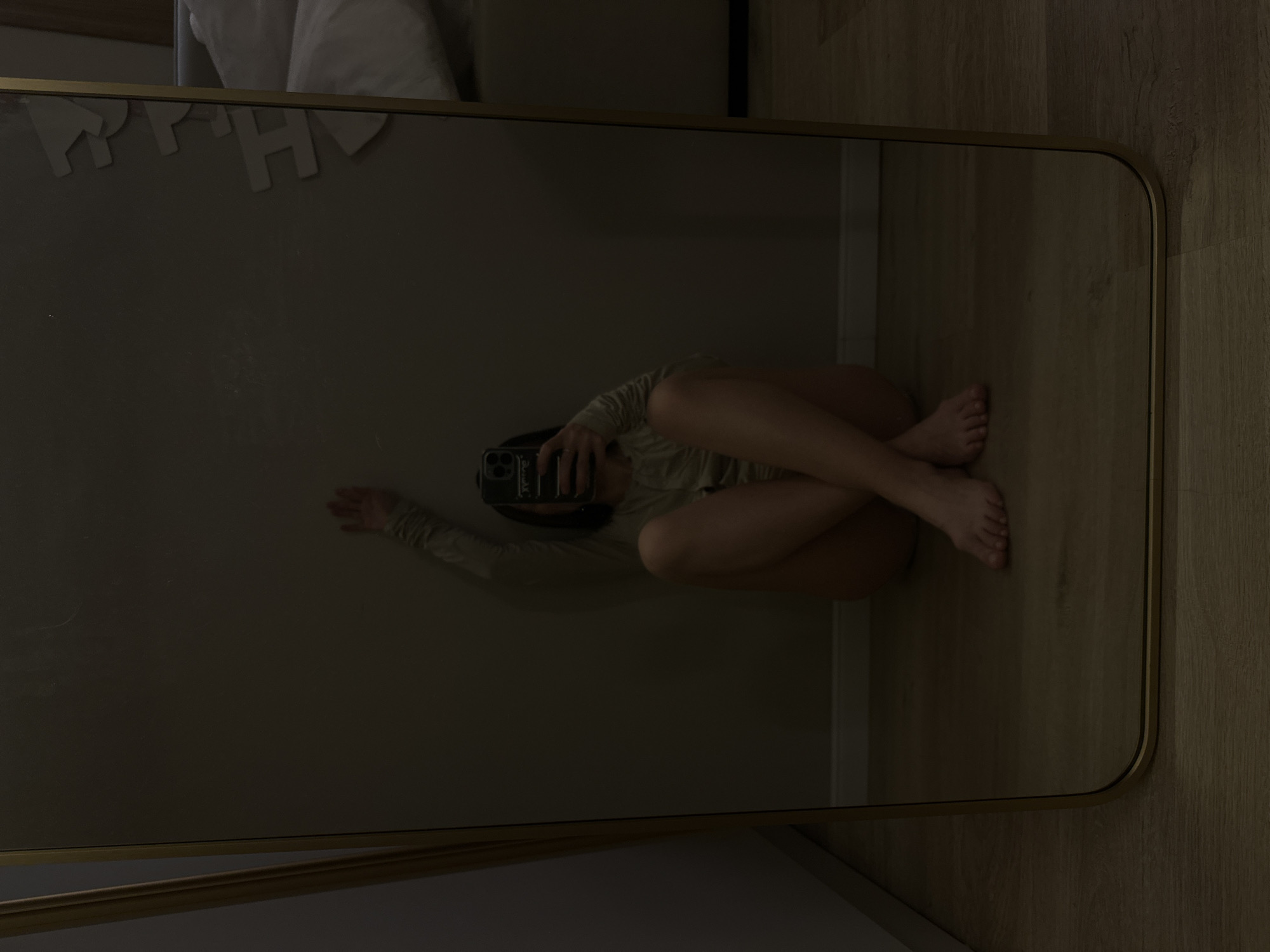 A mirror reflecting a person taking a selfie. The person is sitting on a wooden floor with their legs crossed. The background is a wall with some decorations. The location appears to be an indoor setting. The image does not provide information to determine the city or country of the location.