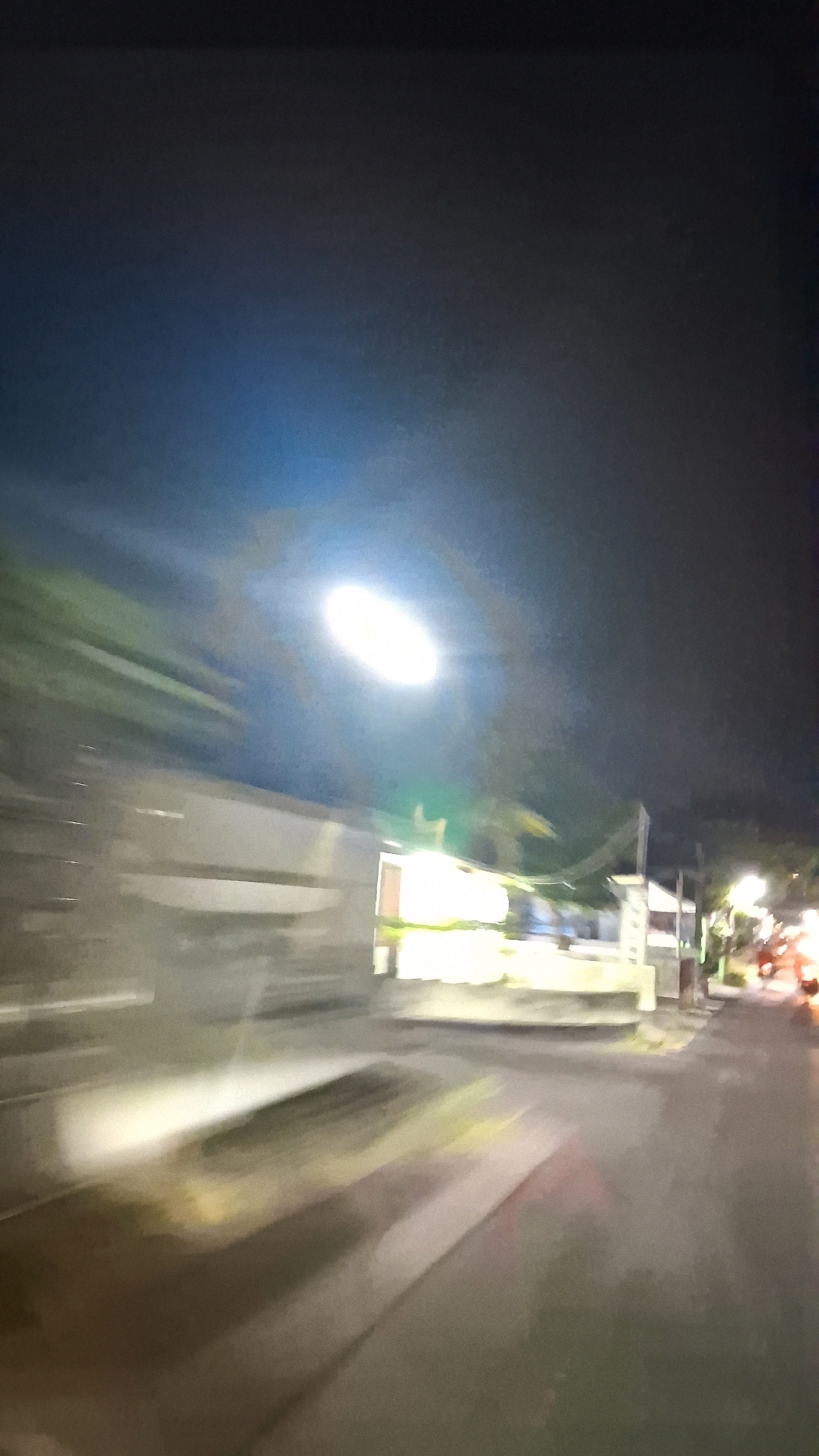 The image depicts a blurred nighttime scene, likely captured from a moving vehicle on a street in Madurejo, Indonesia. A prominent, bright street lamp illuminates the upper left portion of the frame, casting a glare that obscures details in that area. To the left, a blurred structure, possibly a wall or building, runs horizontally. In the mid-ground, indistinct shapes of buildings and vegetation are visible, with some areas brightly lit by artificial light, suggesting shops or residences. The road surface in the foreground is dark and wet, reflecting the streetlights and the movement of the vehicle. Further down the street on the right, blurred lights indicate other vehicles, possibly motorcycles, and streetlights. The overall impression is one of a busy, yet indistinct, urban or suburban street scene at night.