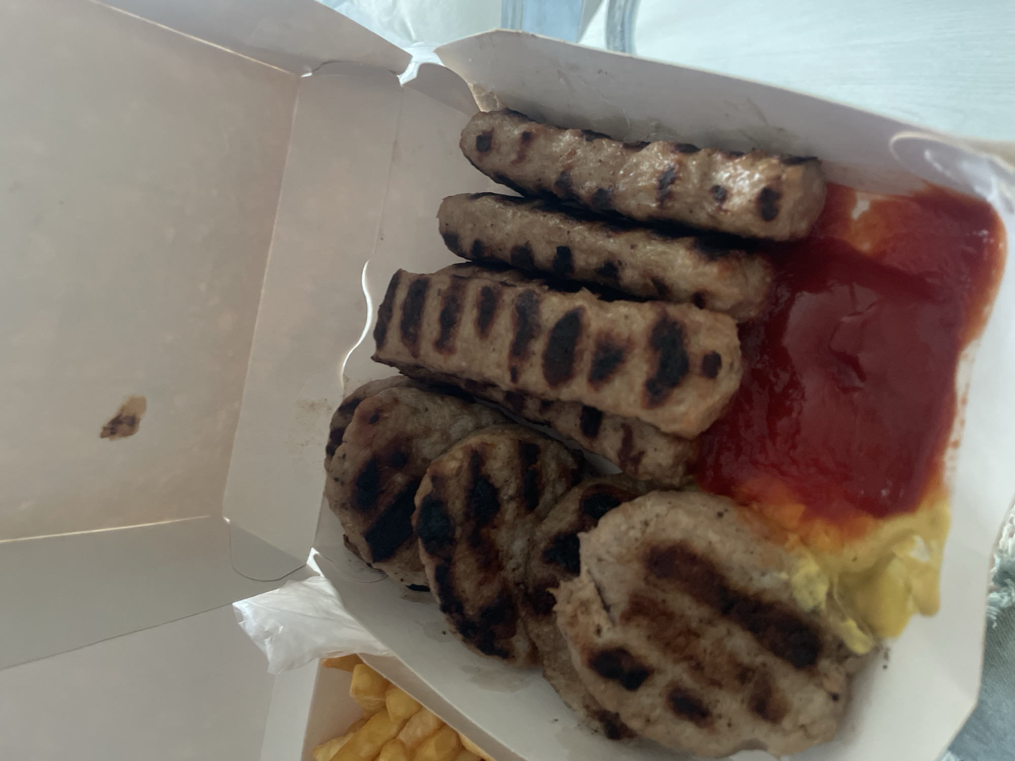 A close-up, high-angle shot captures a take-out meal served in an open white cardboard box, likely indoors during daytime due to the bright lighting. The primary subjects are a selection of grilled meat items, consisting of both elongated, sausage-like pieces and round patties, all featuring distinct char marks from grilling. They are stacked and arranged within the box.
To the right side of the box, generous dollops of red ketchup and yellow mustard are visible, serving as condiments for the meal. In the bottom left corner of the frame, partially visible outside the main box, are golden-brown french fries, suggesting they are an accompaniment to the grilled meats. The overall presentation is casual, typical of a fast-food or street food meal. No people are visible in the image, and there is no discernible text. The scene depicts a meal ready for consumption.