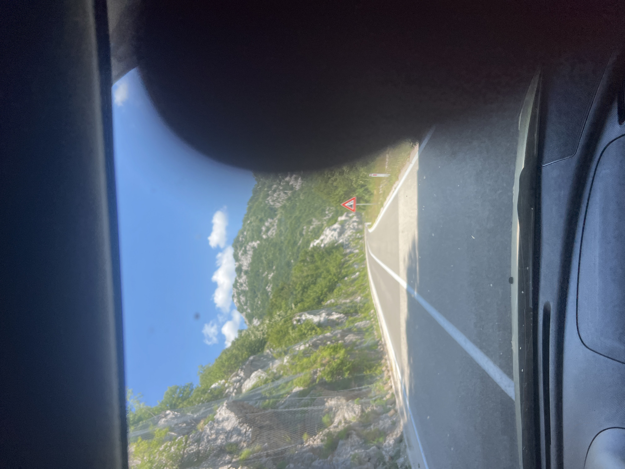 A forward-facing, eye-level shot captures a paved mountain road in Dragalj, Montenegro, on a clear, sunny day. The road curves gently to the right, with a white double line painted down its center, indicating passing is permitted. The terrain is mountainous, with steep, rocky inclines covered in lush green vegetation on the left side of the road. A metal mesh fence runs along the mountainside, presumably to prevent rockfalls. On the right side of the road, a red triangular warning sign with a white border is visible. In the distance, more mountains covered in trees can be seen under a bright blue sky with scattered white clouds. The lower part of the image shows the dashboard and windshield of a vehicle, with a dark, blurry object obscuring the right side of the view. The scene appears to be taken from inside a moving vehicle.