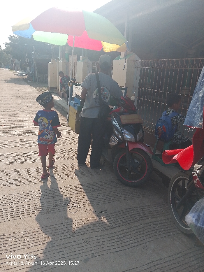The scene is a street in Anjatan, Indonesia (latitude: -6.419, longitude: 107.979). A man in a gray shirt is standing near a parked black motorcycle, which is in the middle of the street. A young boy with a black hat and a blue and red shirt is walking in the opposite direction. A second man is seated behind a red blanket on the sidewalk, which is lined with a metal fence. A young boy in a blue shirt is seated next to the red blanket. A colorful umbrella is positioned to the left of the scene. The ground is paved with concrete and has a textured surface. The scene is lit with a bright, sunny light. The photograph was taken at 15:27 on April 16, 2025.