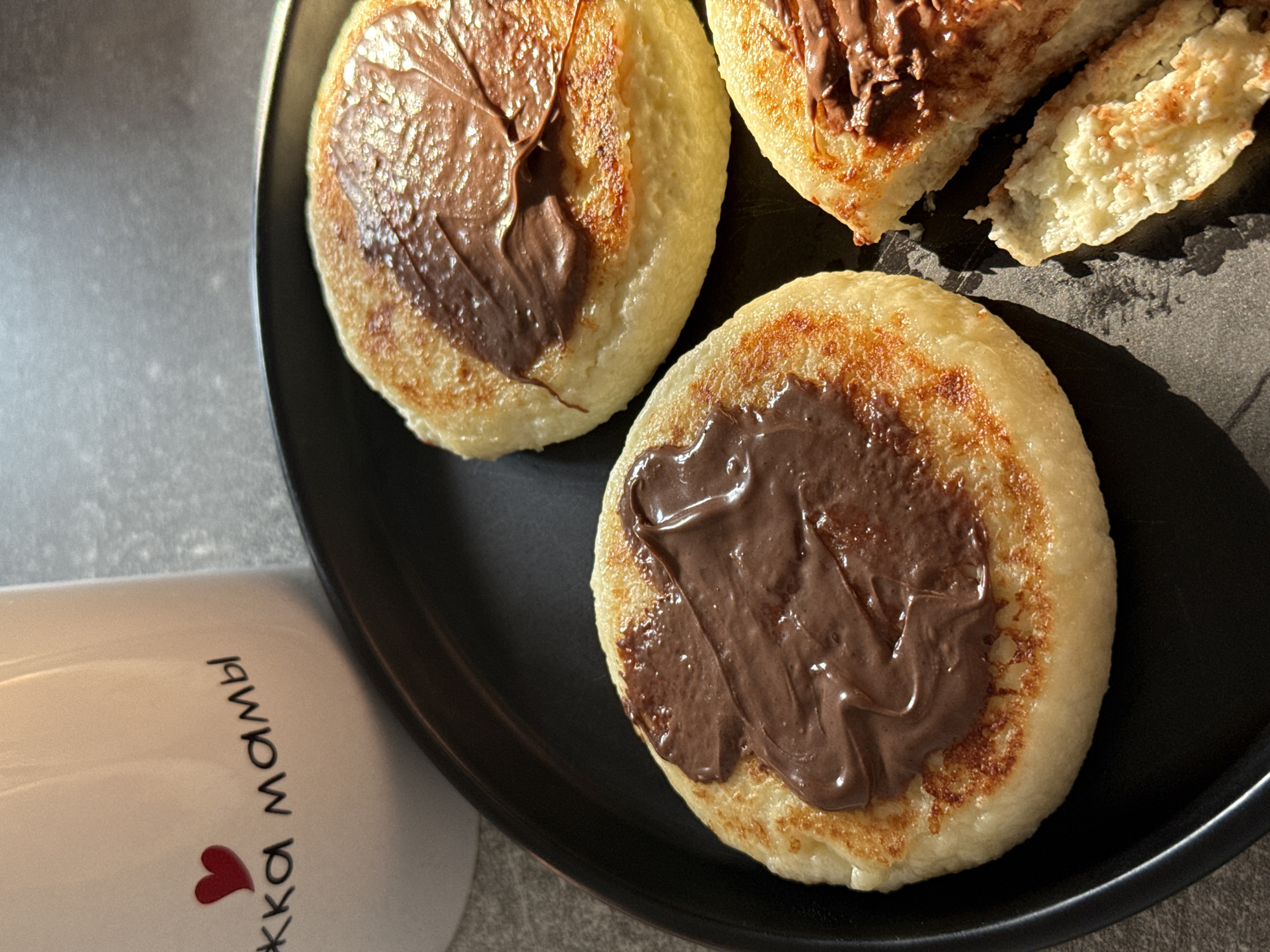 A black plate holds three syrniki (fried cottage cheese pancakes) on a gray countertop. The syrniki are golden brown and topped with chocolate spread. A white mug with a red heart and Russian text is partially visible in the upper left corner. The scene suggests a domestic setting. There is no indication of the city of Lyubertsy, Russia.