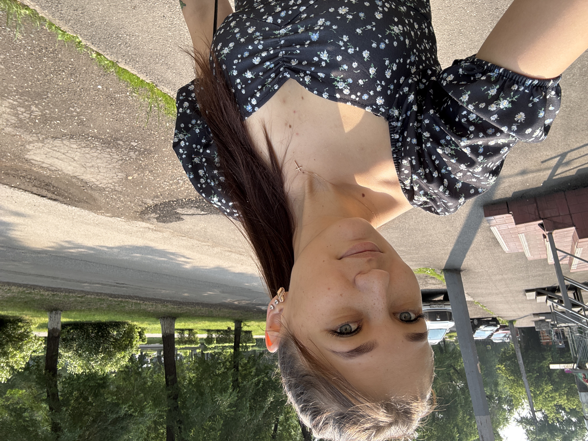In Novokuznetsk, Russia, a young woman is captured in a selfie outdoors on a sunny day. She is wearing a black dress with a floral pattern and a delicate cross necklace. Her hair is pulled back in a ponytail, and she has a light makeup application. In the background, a paved walkway leads to a building with stairs, and a street with parked cars is visible. Trees and green bushes line the sidewalk, indicating a residential or park-like area within the city. The lighting suggests it is daytime.