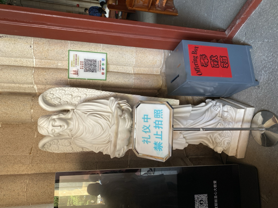 This vertical shot, rotated 90 degrees clockwise, captures an indoor scene likely within a church or cathedral, specifically identified as the Guangzhou Sacred Heart Cathedral.
In the foreground, a large white statue of an angel with detailed wings stands, holding an object in its arms. Attached to the angel's base is a white sign with blue Chinese characters stating, "礼仪中禁止拍照" (Lǐyí zhōng jìnzhǐ pāizhào), meaning "No photography during ceremony/ritual."
To the right of the angel statue, a grey "Offering Box" is visible. It features red labels with white text that read "Offering Box" in English and "奉獻箱" (Fèngxiàn xiāng), meaning "Offering Box," in Chinese.
On the stone wall behind the angel, a green sign with a QR code and Chinese text is displayed. The text includes a "温馨提示" (Wēnxīn Tíshì - Warm reminder) advising visitors to "建议参观圣心大教堂 请勿大声喧哗" (Jiànyì cānguān shèngxīn dà jiàotáng qǐng wù dàshēng xuānhuá), which translates to "When visiting the Sacred Heart Cathedral, please do not make loud noises." Below the QR code, "广州石室圣心大教堂" (Guǎngzhōu Shíshì Shèngxīn Dàjiàotáng), or "Guangzhou Sacred Heart Cathedral," is written, along with contact numbers (e.g., 020-83336711) and social media icons.
In the bottom left, a reflection in a dark, possibly glass, surface shows a partial view of what appears to be a person and outdoor greenery. Another QR code and the text "聖心大教堂" (Shèngxīn Dàjiàotáng - Sacred Heart Cathedral) are faintly visible on this surface. In the background, wooden pews and a person are partially visible, reinforcing the church setting. The lighting is bright, suggesting daytime.