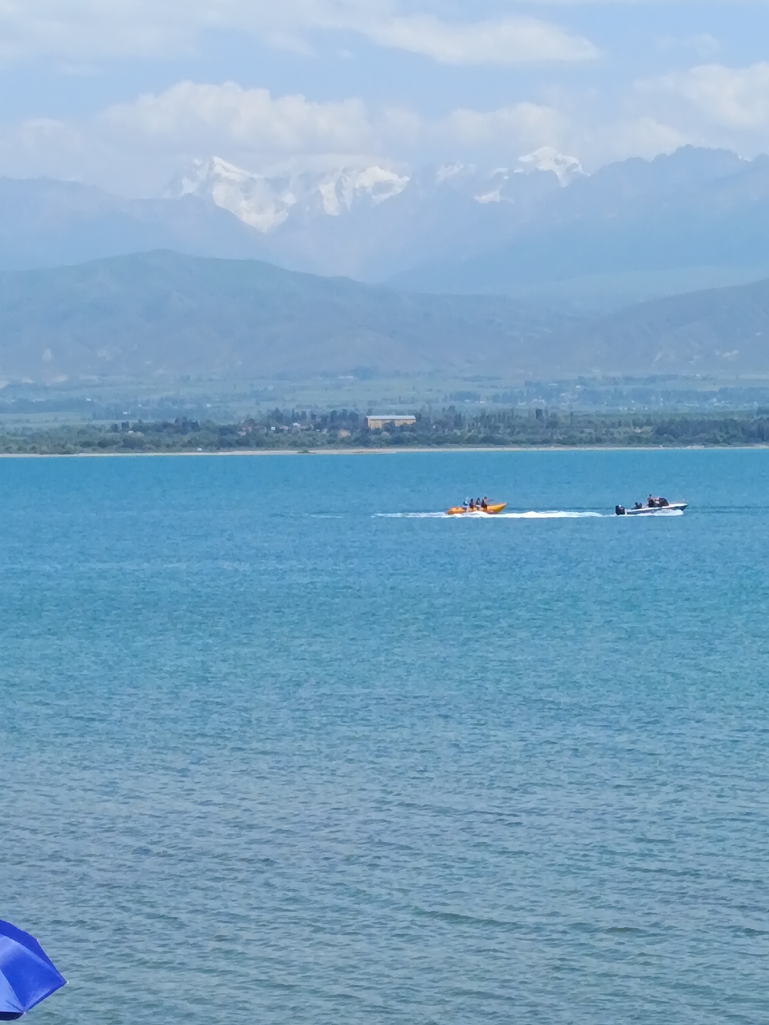 An eye-level, medium shot captures a serene wide body of bright blue water with a gently rippling surface under a partly cloudy sky. In the mid-ground, two motorboats are actively moving across the water. The boat on the left is yellow and appears to be a towable inflatable, with several people riding on it. It is creating a white wake behind it. To the right of the yellow boat, a white motorboat with a dark top is also speeding across the water, with at least two individuals visible on board. The boats are heading in the same direction, leaving trails in the water. The background features a long, low stretch of green land with trees and buildings, suggesting a populated shoreline. Beyond the land, imposing mountains rise, their lower slopes a muted green and blue, transitioning to snow-capped peaks that are brightly lit by the sun. The sky above the mountains is a pale blue with scattered white clouds. In the lower-left corner of the frame, a portion of a bright blue umbrella is visible, suggesting the presence of a beach or viewing area. The overall impression is a scenic, active waterscape with a dramatic mountain backdrop, likely taken in or near Kyzyl-Suu, Kyrgyzstan, given the context of the snowy peaks and a large body of water.