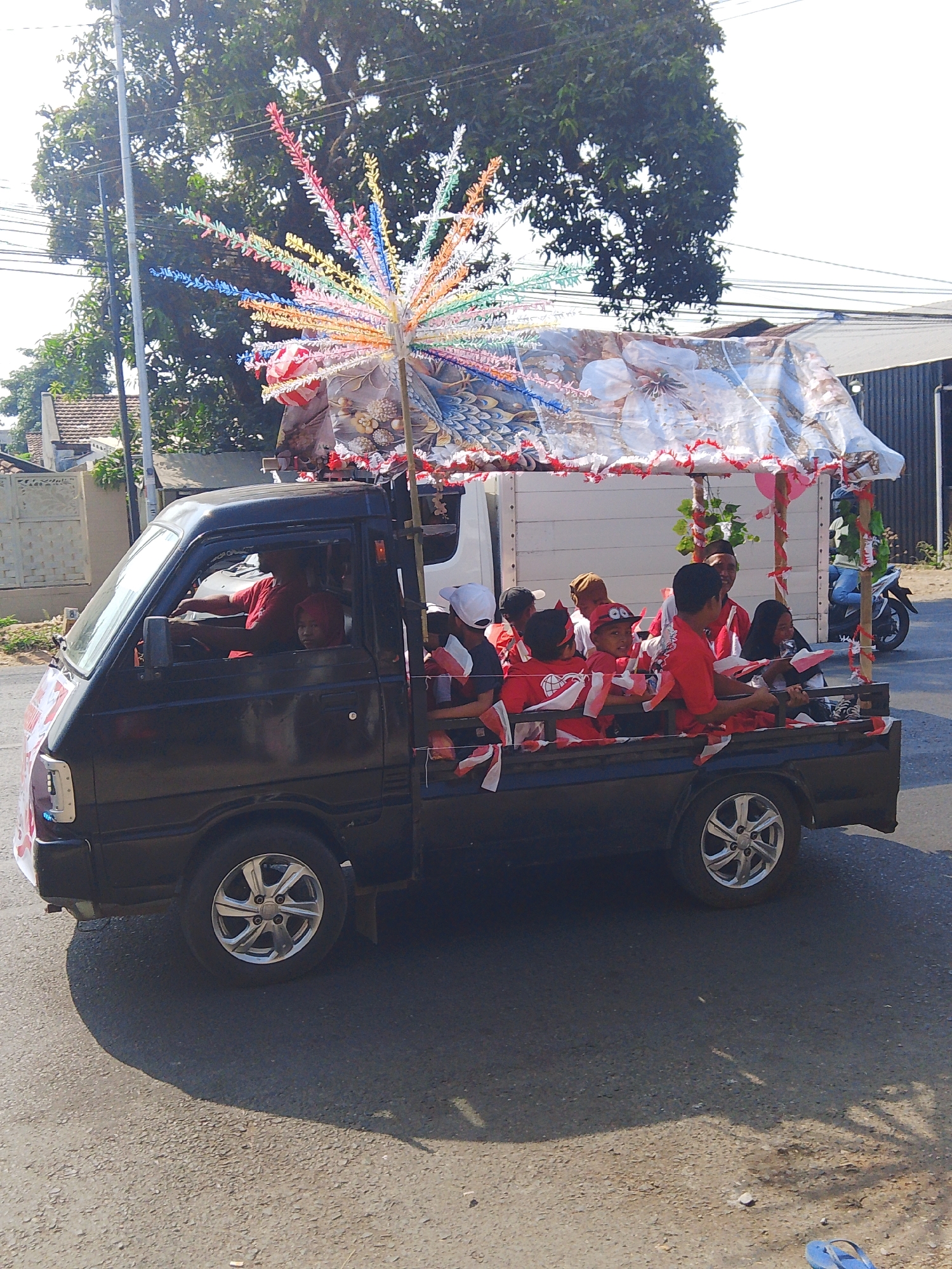 A black pickup truck, decorated with a large, colorful, fan-like structure made of plastic strips and fabric, is driving down a paved street in Kalinyamatan, Indonesia. The truck is carrying several people, mostly children, who are wearing red shirts and are holding small Indonesian flags. Some of the passengers are holding onto the sides of the truck, while others are seated. The truck has a canopy made of fabric that is also decorated with flags and other ornaments. A white building with a tiled roof is visible in the background, along with a large tree and power lines. The sky is bright and clear. The scene appears to be a parade or a celebration.