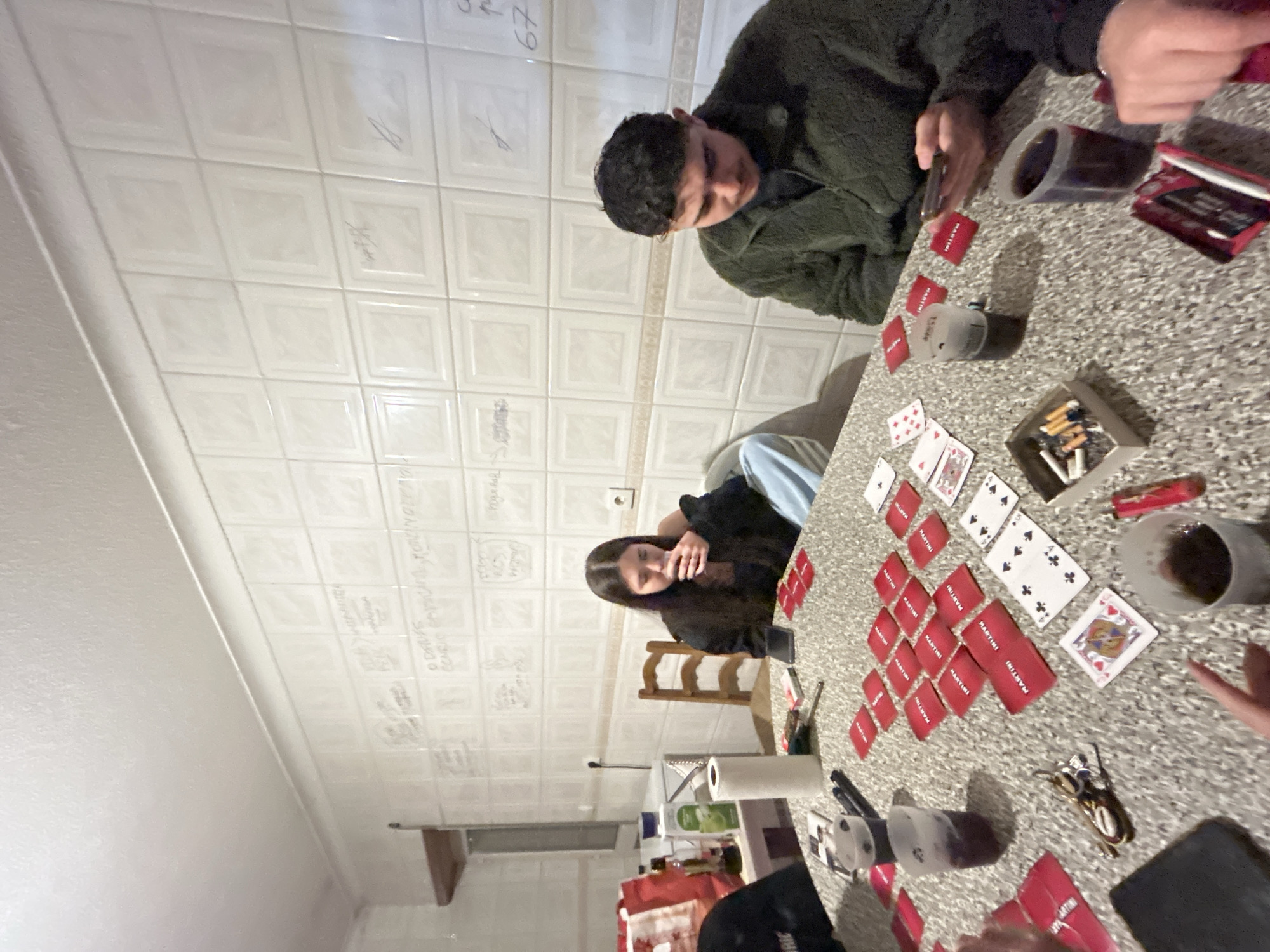 The image depicts an indoor, informal social gathering in Bragança, Portugal, characterized by its distinctive tiled walls. These off-white, square ceramic tiles are extensively covered with handwritten inscriptions, signatures, and simple drawings in various dark markers, primarily in Portuguese (e.g., "O DINIS COMEU SAPUCAIAS," "67"). This style of inscribed walls is characteristic of informal taverns or student gathering places in the city. The ceiling is plain and textured white.
In the foreground, a speckled light-grey table is the central point of activity, hosting a card game involving at least four individuals. The game setup includes numerous red-backed "MARTINI" branded cards, some face down, others forming stacks. Several standard playing cards, such as the Ace of Spades, 4 of Spades, 8 of Clubs, 10 of Spades, 9 of Clubs, and Queen of Hearts, are visible face-up.
Multiple clear plastic cups containing a dark liquid are distributed around the table. An improvised ashtray, a small cardboard box, holds cigarette butts and loose tobacco/cigarettes, with a red lighter nearby. Personal items include a set of keys with a keychain, and at least two smartphones; one rests on the table in front of a female subject, and another is actively held by a male subject. A dark, rectangular object, possibly another smartphone or tablet, is partially visible in the bottom left. A roll of white paper towels is also on the table.
Of the people present, a female individual with long, dark hair is seated center-left, wearing a dark top and light blue pants. She is seen bringing a clear plastic cup to her lips. To her right, a male individual with dark, curly hair is seated, wearing a dark green textured jacket over a dark shirt. He is looking down at a smartphone held in both hands. Portions of other individuals are visible: a dark sleeve and a leg in light blue pants are on the far left, and a hand holding a cup is in the bottom right.
In the background to the left, a surface holds a dark red bag, multiple bottles (one dark-colored, possibly olive oil, and another containing a light green liquid), and a white roll of paper towels. A light-colored wooden chair is also visible. The overall scene suggests a casual, late-night social interaction among friends or acquaintances.