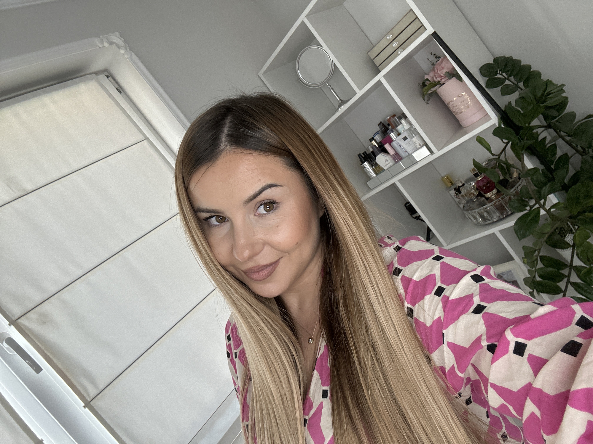 A medium close-up, eye-level selfie captures a woman indoors, in what appears to be a personal living space in Asenovgrad, Bulgaria. She has long, straight, blonde hair with visible darker roots, parted down the middle, flowing over her shoulders and in front of her. Her face is turned slightly to the right, and she looks directly at the viewer with a gentle, subtle smile and warm brown eyes. She wears light makeup, enhancing her features, and a thin golden necklace with a small, round pendant is visible on her chest.
She is dressed in a vibrant top or dress featuring a repeating geometric pattern of fuchsia pink, white, and black shapes.
The background reveals a bright, modern room. To her left, a white window or door frame is partially visible, indicating a source of natural light. Behind her, a white open-shelf unit is filled with various personal items. On the shelves, a silver vanity mirror, small decorative boxes (one with jewel-like handles), and numerous perfume bottles and cosmetic items are neatly arranged, some on a mirrored tray. A pink cylindrical vase holding artificial pink flowers is also present. A lush green plant, possibly a ZZ plant, is partially visible on the far right, adding a touch of greenery to the clean aesthetic. The lighting suggests it is daytime. No discernible text is clearly readable on any of the objects.