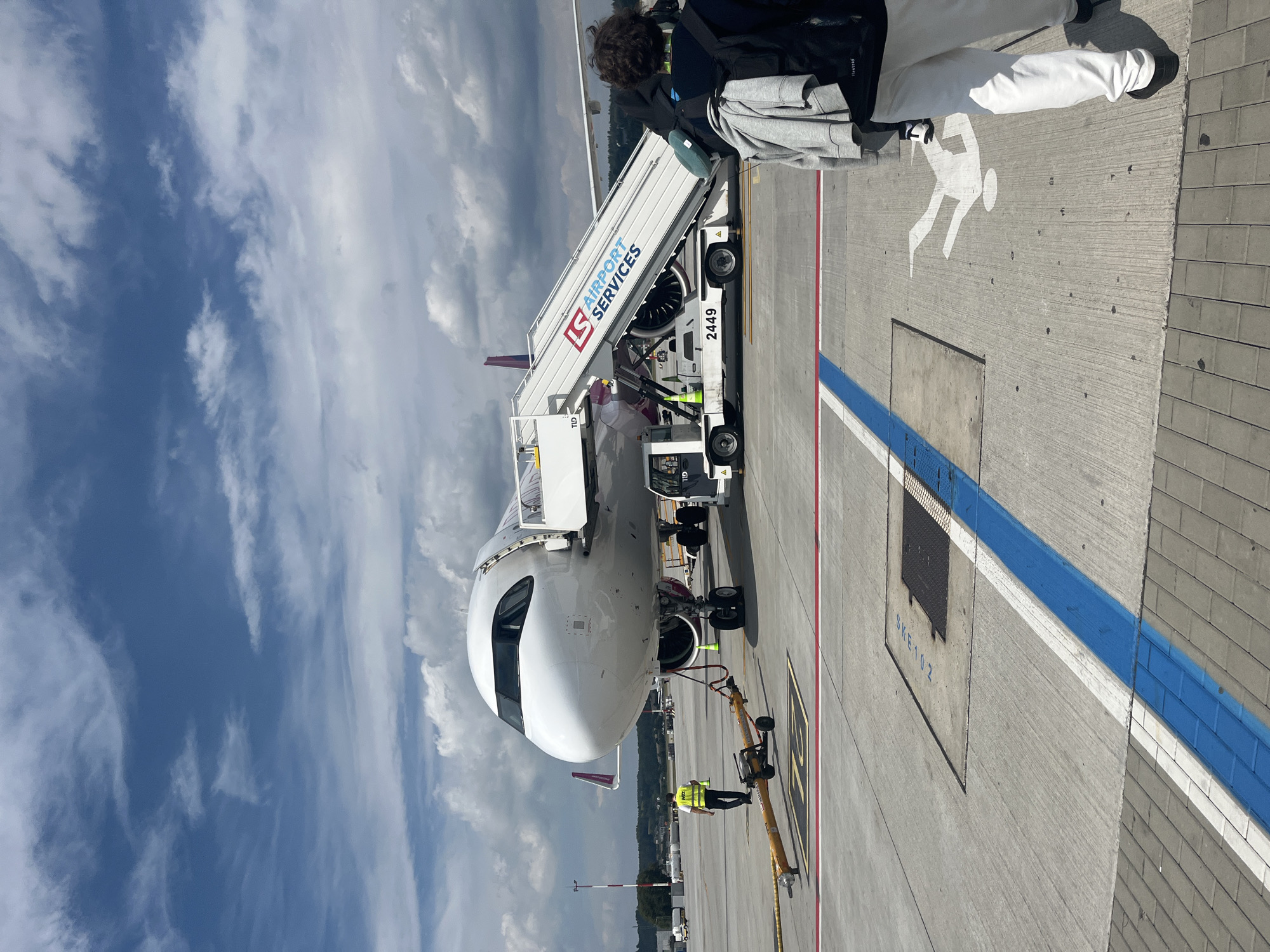 An airplane is parked on the tarmac at Balice, Poland, a town near Krakow. The aircraft, a white Airbus A320 with purple accents, is attended by ground crew and a mobile stairway. A person in a high-visibility vest walks near the front of the plane, beside a tow bar. Passengers are disembarking or boarding via the stairs, which bear the "LS AIRPORT SERVICES" logo. The ground is marked with yellow and blue lines, and a white pedestrian symbol is painted on the concrete. The sky is partly cloudy, with a mix of blue sky and white cumulus clouds. In the background, trees and buildings are visible, suggesting the airport is close to a developed area.