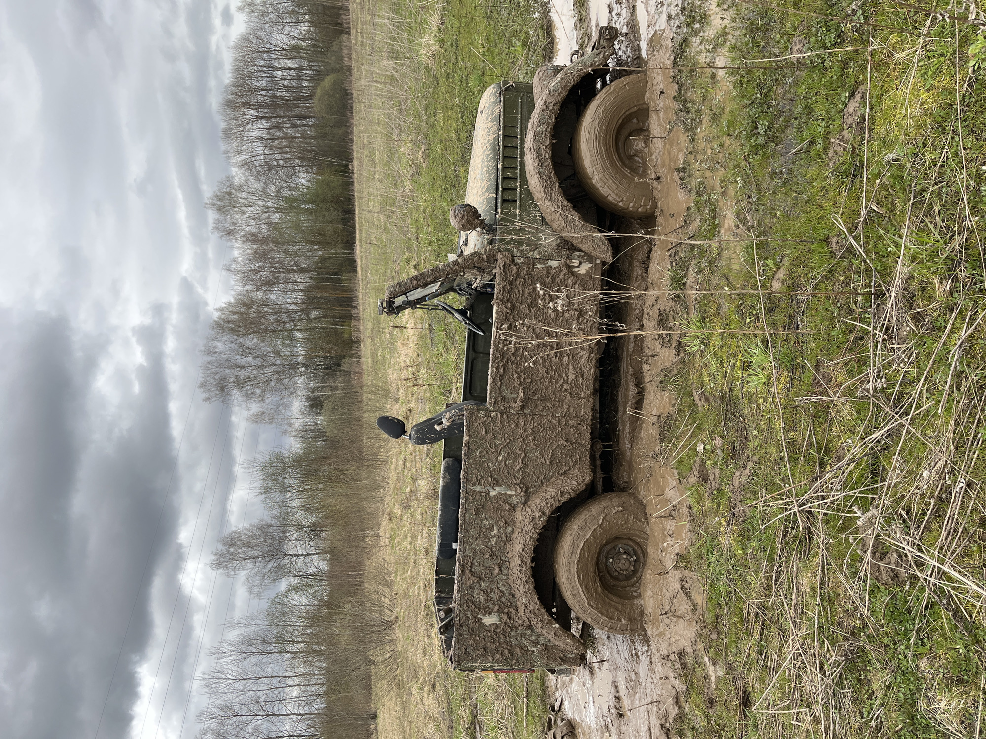 The image depicts a mud-covered military vehicle stuck in a muddy field. The location is presumably in the vicinity of Раздолье, Russia. The vehicle is an open-topped utility vehicle, possibly a military jeep or similar model, covered in thick mud from tires to windshield. The vehicle is on a muddy path or track within a field. The tires are deeply sunk into the mud. The surrounding environment consists of fields, some with sparse green grass, and a distant treeline under a cloudy sky. The trees appear bare, suggesting it might be early spring or late autumn. Power lines are visible on the left side of the image.