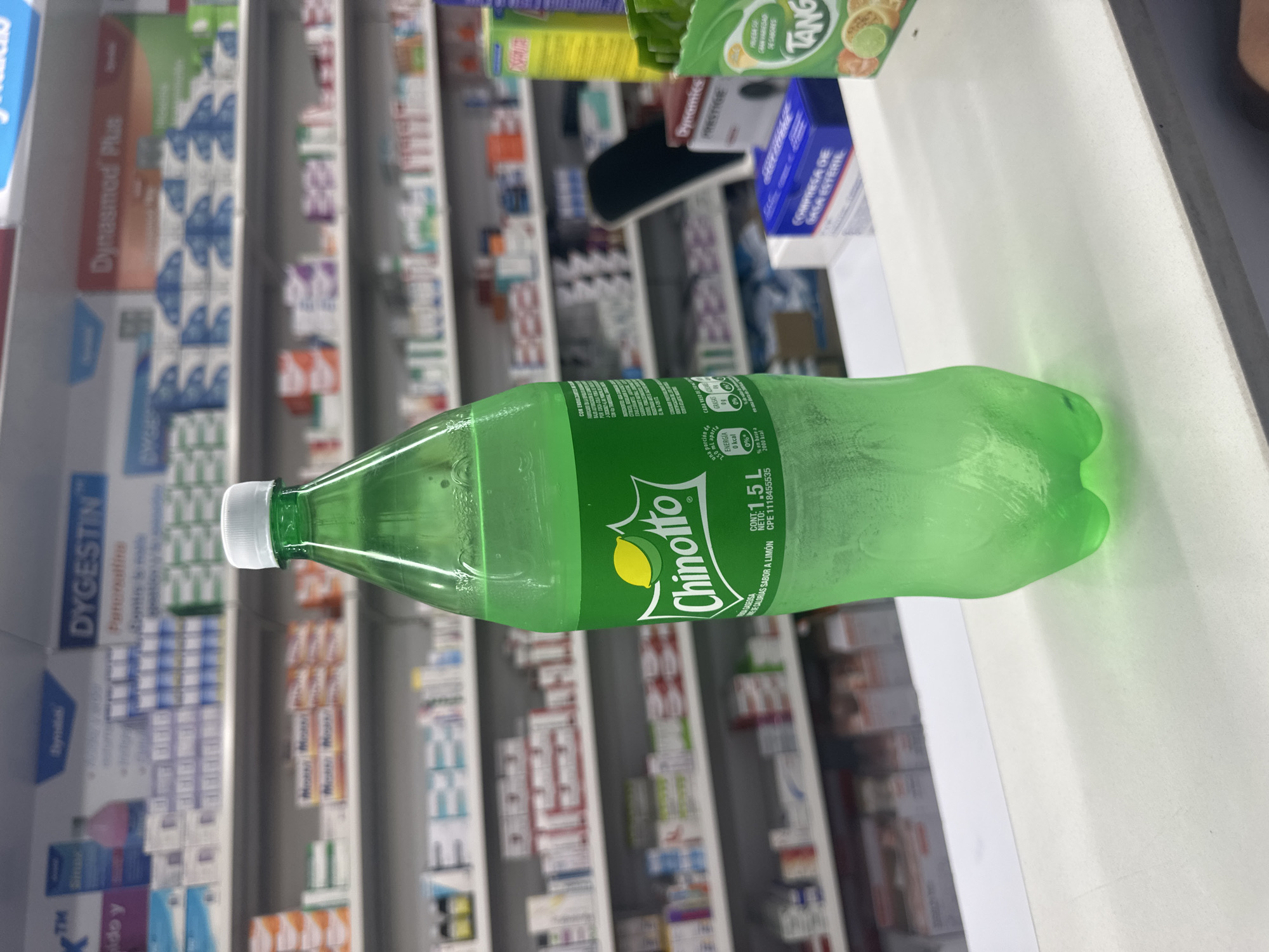 A 1.5L bottle of Chinotto lemon-flavored soda lies horizontally on a white surface in what appears to be a pharmacy in Parroquia Ocumare, Venezuela. Rows of pharmaceuticals in boxes and packages fill shelves in the background, including Dynasmed Plus and Dygestin. Additional products, including Tang flavored drink mix and "Dynamics Prestige" brand items, are partially visible on the surface near the bottle. The Chinotto bottle is green with a white cap.
