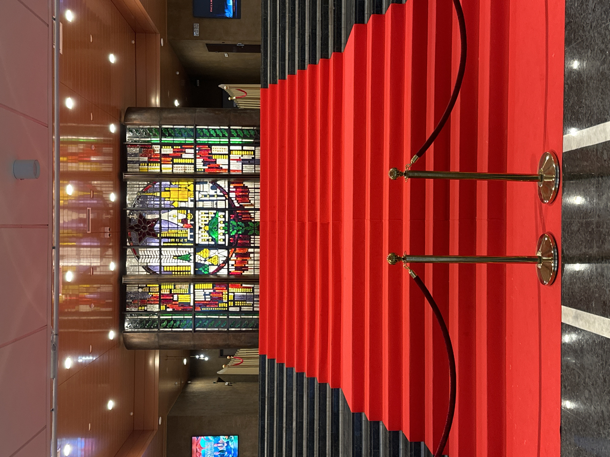 This image captures the grand interior of the Palace of the Republic, located on October Square in Minsk, Belarus. The scene features a wide central staircase meticulously covered by a vibrant red carpet, extending from the foreground to a landing directly beneath a large, colorful stained-glass window. Two golden stanchions with dark red velvet ropes are positioned in the immediate foreground, delineating the entrance to the red carpet. The sections of the staircase to the left and right of the carpet are composed of dark material with lighter-colored horizontal lines.
The prominent stained-glass window in the background features a red five-pointed star at its top center, with a hammer and sickle emblem directly below it. The central portion of the window depicts urban architecture rendered in various hues of yellow, green, red, and blue. Flanking panels display abstract or geometric patterns in similar color schemes. The window is framed by a dark, rounded column on its right side.
The ceiling above is constructed from light-toned panels with embedded circular recessed lights, which cast reflections on the glossy ceiling surface. On the left wall, a digital display screen shows an image of two individuals and the Cyrillic text "УМНЫЕ СОЗДАНИЯ" (Smart Creations). On the right wall, a digital clock displays "20:30" above a screen exhibiting the Cyrillic word "ПАССАЖ" (Passage). A dark doorway is visible further along the right wall. The floor in the foreground, beneath the stanchions, is dark, reflective, and patterned with lighter streaks. No people are present in the depicted scene.