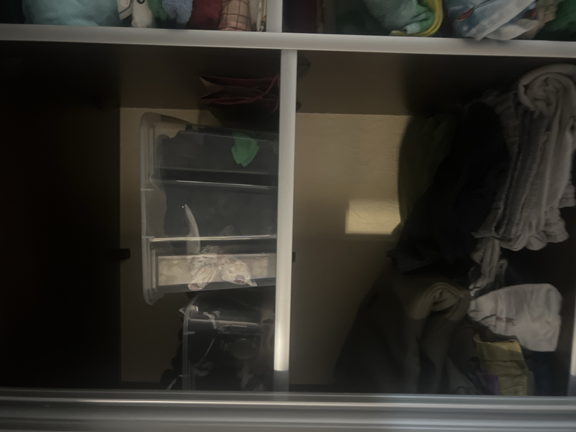 The image depicts the interior of a closet, showcasing two shelves filled with various items. The top shelf holds clear plastic storage bins containing folded clothing and other items. One bin is partially obscured, but its contents appear to be dark fabric. Another bin contains lighter-colored items with a patterned design. To the right of these bins, a red handbag or pouch is visible.
The bottom shelf is filled with a large stack of folded textiles, likely blankets or towels, in various shades of gray, beige, and dark blue. Some items are rolled, while others are neatly folded.
The closet appears to be located in a residential setting, possibly a home or apartment. The lighting is dim, suggesting the closet door is closed or the ambient light is low. There is no indication of the specific location within the city of Surgut, Russia, nor are there any people or discernible actions taking place. The scene is static and focuses on the organization and contents of the closet.