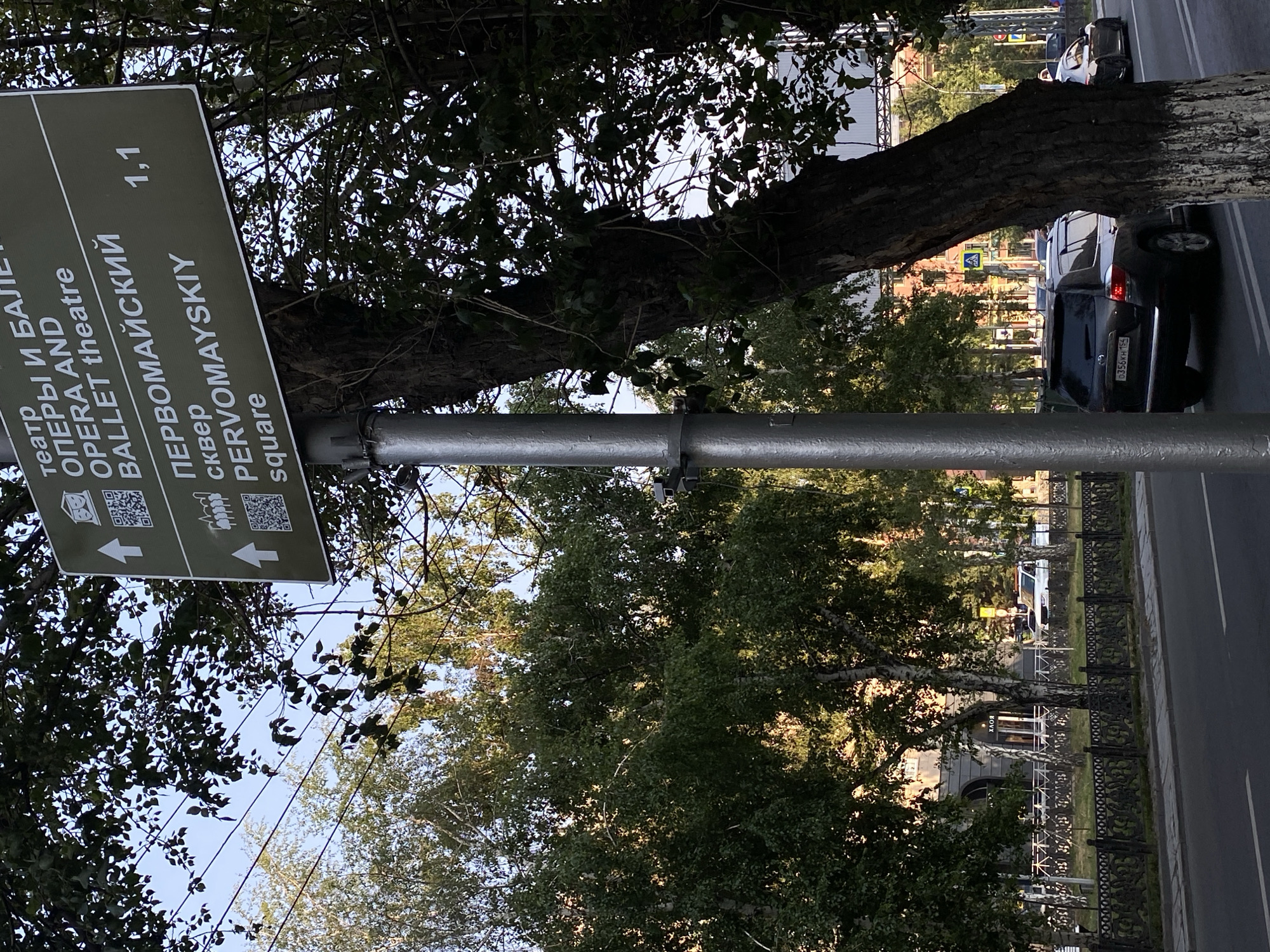 This image depicts a street scene in Novosibirsk, Russia. A tall, slender metal pole stands in the foreground, supporting a green directional sign. The sign displays text in Russian and English, indicating "ТЕАТР ОПЕРЫ И БАЛЕТ" (OPERA AND BALLET THEATRE) with an arrow pointing left and a distance of "1,1". Below this, it reads "ПЕРВОМАЙСКИЙ сквер" (PERVOMAYSKIY square) also with a left-pointing arrow. The sign includes QR codes next to each direction. The background features a street with cars, trees, and buildings. A dark-colored SUV is parked on the right side of the street, and another car is visible further down. Trees with lush green foliage line the sidewalk, and a decorative metal fence is present. The lighting suggests it is daytime, likely late afternoon or early morning due to the angle of the sun creating shadows.