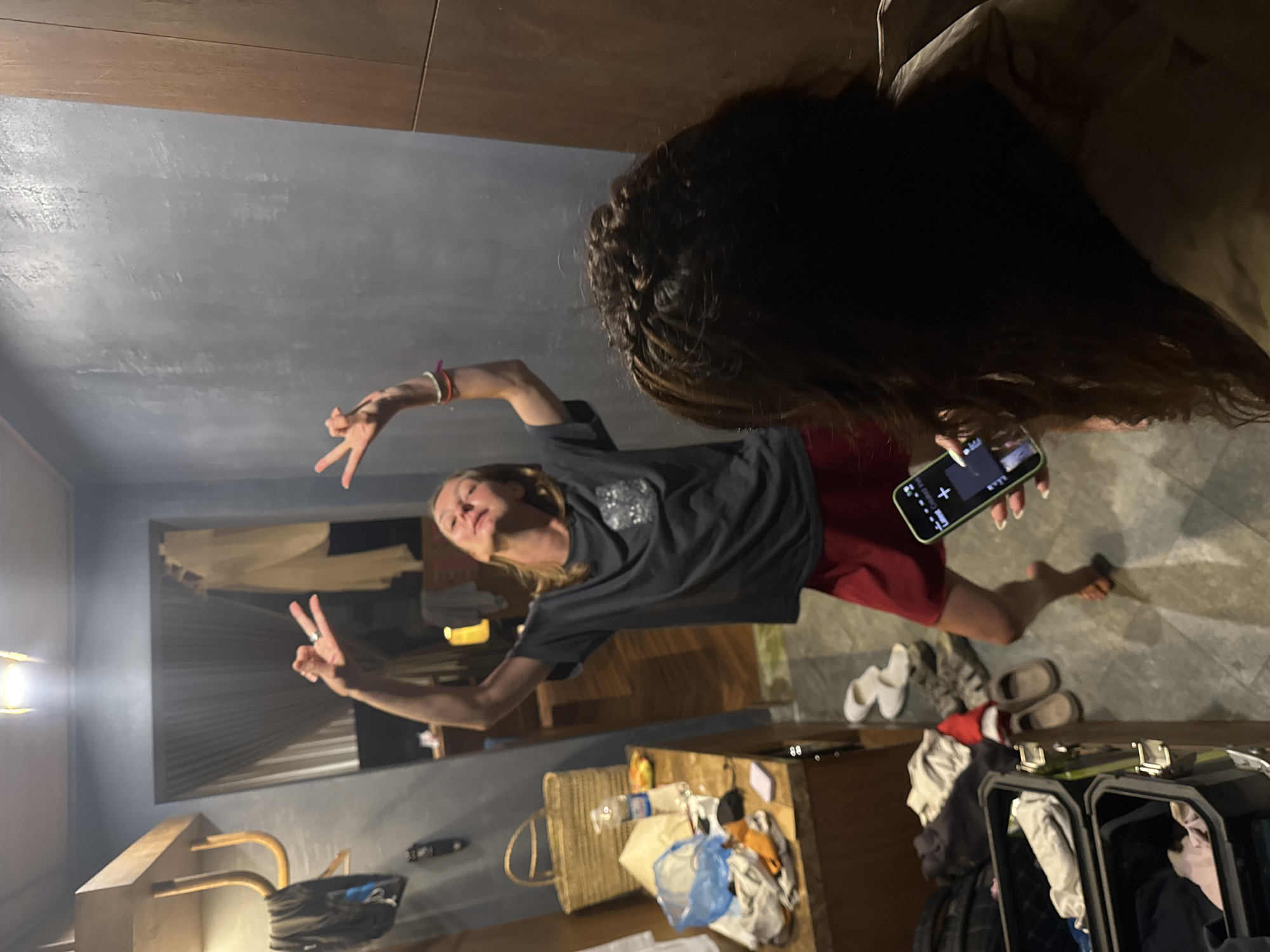 This indoor scene, set in Mù Cang Chải, Vietnam, features two individuals within a room characterized by gray, textured walls and dark floor tiling.
In the mid-ground, a light-skinned female with light brown hair and wearing a dark gray t-shirt and red shorts, stands barefoot. Her head is tilted back with eyes closed, and both arms are raised, hands forming peace signs. Colorful bracelets adorn her right wrist, and a ring is visible on her left index finger.
In the foreground, partially visible from the back, a person with dark, braided hair and wearing a light brown garment holds a light green smartphone, pointed towards the first individual. The phone's screen displays a dark interface.
The room's left side features a wooden wall-mounted shelf with hangers, from which dark clothing hangs. Below this, a wooden counter is cluttered with a woven straw basket, a partially full plastic water bottle, yellow snack packaging, folded clothing, a blue plastic bag, a black remote control, and a small white object. On the floor below the counter, two open black luggage cases contain various garments. Additional footwear, including white slippers, brown slip-on shoes, and dark sandals, is visible on the floor, alongside a red Santa hat. A bright ceiling light fixture is centered at the top of the image. Through a doorway or opening in the background, a window with light-colored curtains and another ambient light source are visible.