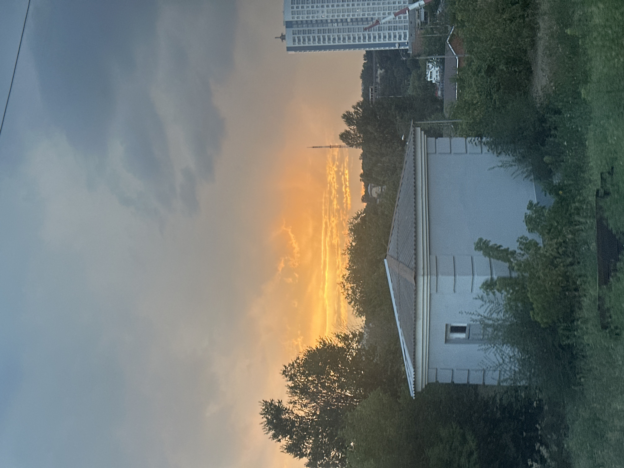 A wide shot captures a sunset over a suburban area in Rostov-on-Don, Russia. The sky is a dramatic mix of dark, brooding clouds and bright, fiery orange hues from the setting sun, with rays of light piercing through. In the foreground, a small, octagonal, light blue building with a gray, corrugated roof and a single small window is visible, partially obscured by lush green foliage and bushes. To the right, a tall, modern, white apartment building dominates the skyline, with a red and white crane positioned in front of it. A radio tower stands in the distance, beyond a dense line of trees that separate the foreground from the cityscape. The overall scene suggests the transition from day to night in a developed, yet green, urban environment.