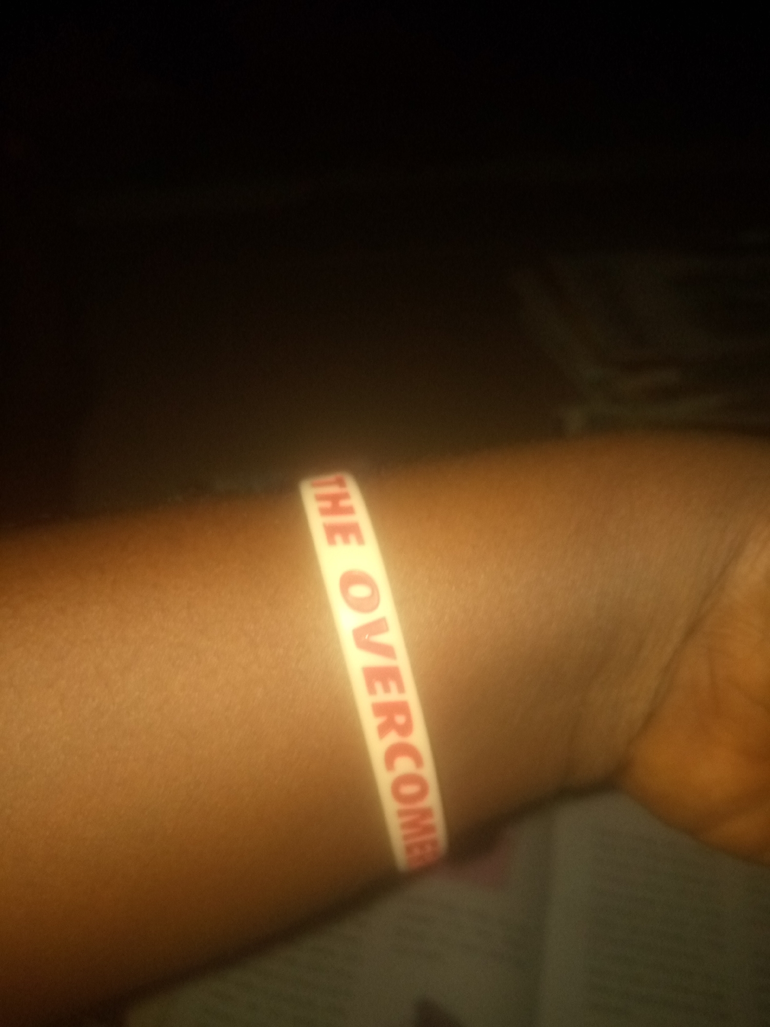 A close-up, dimly lit, horizontal, eye-level shot captures a person's arm and wrist, adorned with a white rubber bracelet featuring red lettering. The bracelet is partially visible, with the words "THE OVERCOME" clearly legible. The arm appears to be tanned, and the skin texture is visible, with subtle pores and hairs. The background is dark and out of focus, suggesting an indoor environment, possibly a room with low lighting. No specific urban or environmental details beyond the arm and bracelet are discernible.