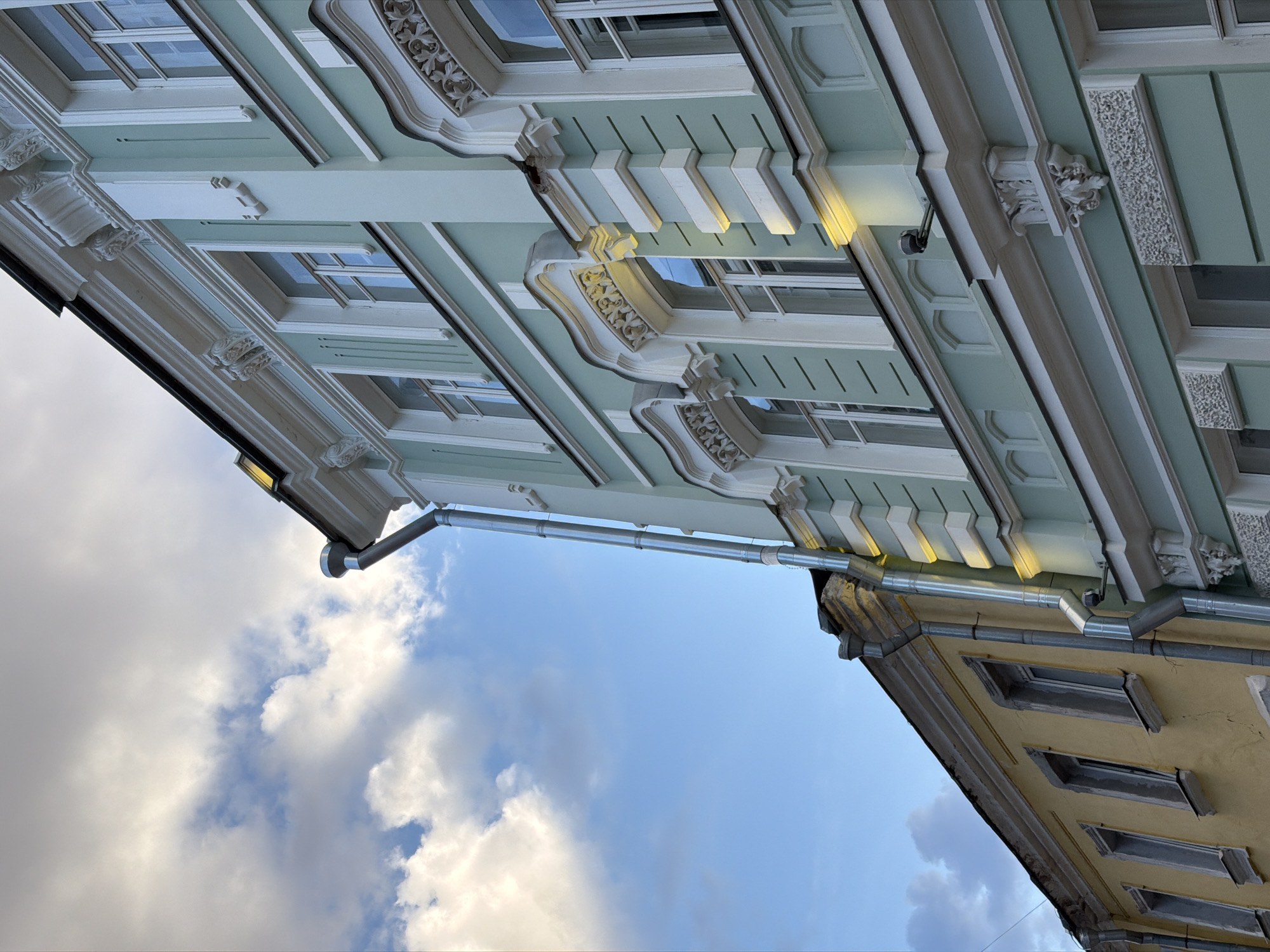 The image captures an upward view of two buildings in Moscow, Russia, against a backdrop of a partly cloudy sky. The building on the left is a pale green color, featuring ornate architectural details, including window frames, moldings, and decorative relief work. The building on the right is yellow and displays similar but less elaborate architectural features. A metal downspout connects the two buildings. The cloudy sky creates a soft, diffused lighting effect on the building facades.