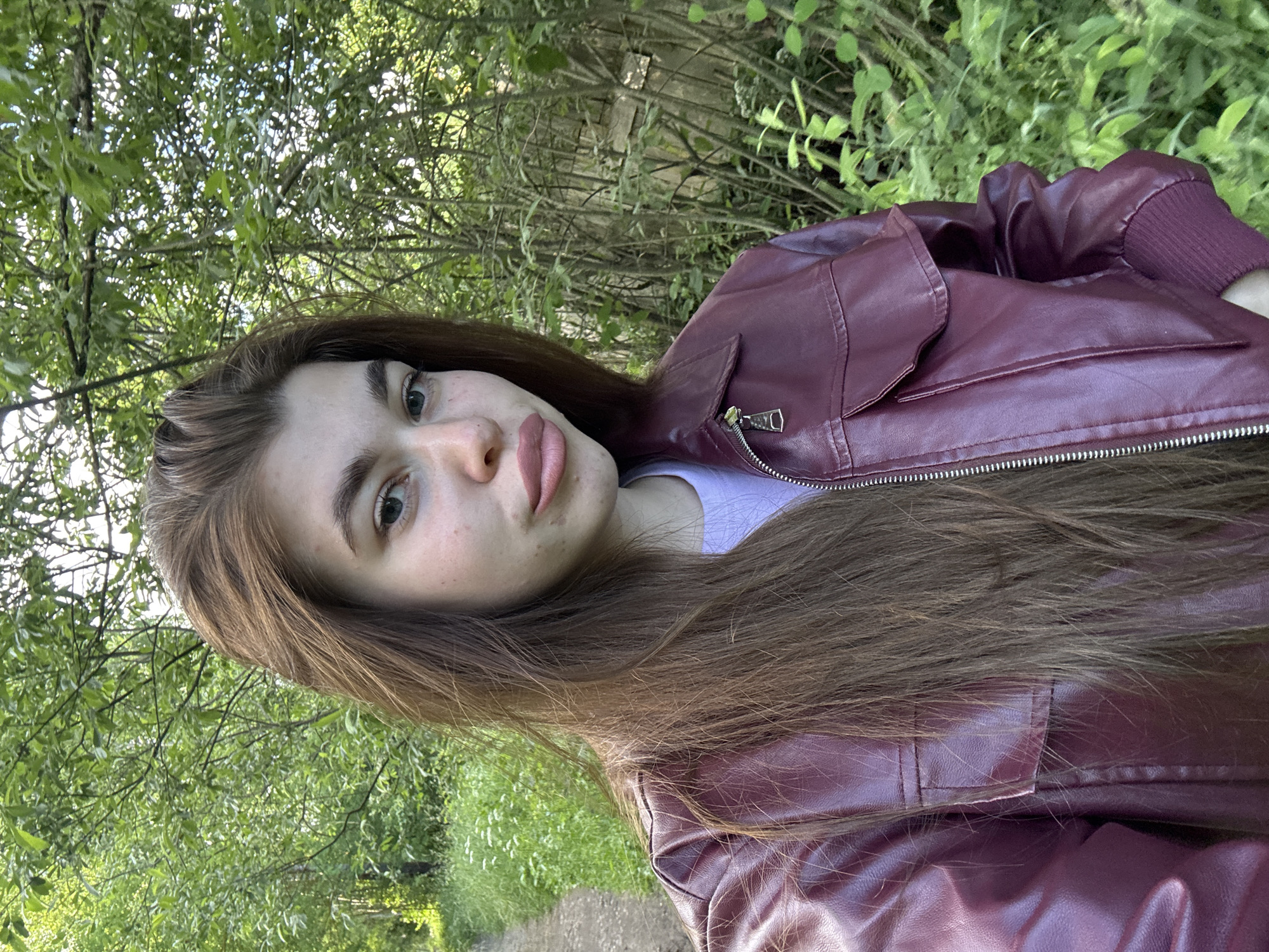 A young woman with long, brown hair and a maroon faux leather jacket poses for a selfie outdoors. She has light skin with freckles and is wearing a light purple t-shirt underneath her jacket. Her lips are slightly parted and she is looking towards the camera. The background consists of lush green trees and foliage, suggesting a natural, possibly rural or park-like setting. The path to her left indicates a walking trail. The location is identified as Мещёры, Russia.