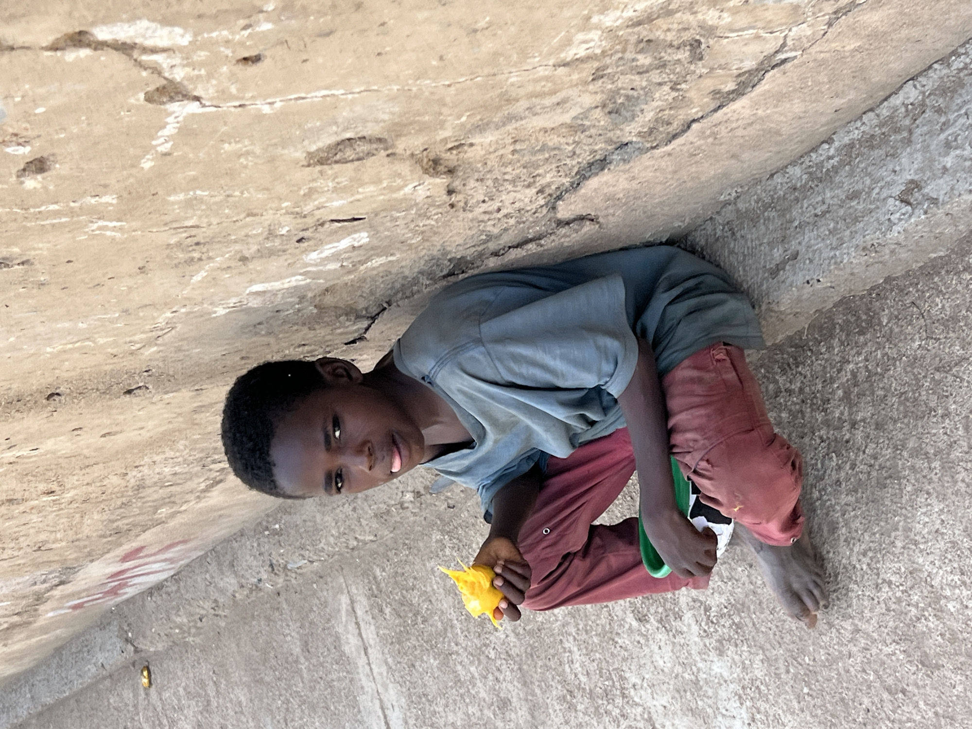 A young Black boy is crouched in a corner formed by a concrete structure. The scene appears to be set in an urban location, possibly Jos, Nigeria, given the context provided. He is wearing a light blue t-shirt and reddish-brown pants. He is holding a piece of yellow food, perhaps fruit, in his right hand. He also has a small green and white object in his left hand. The surface around him is cracked concrete. The boy is looking at the camera. He is barefoot.