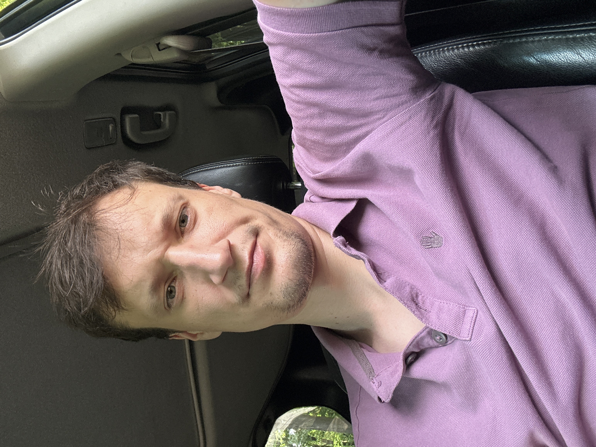 A man is sitting in the driver's seat of a car, looking at the camera. He has short, dark brown hair and a light complexion. He is wearing a light purple polo shirt. The interior of the car is visible behind him, including a black leather seat and part of the car's roof. Outside the car, through a window on the left, trees and greenery are visible, suggesting an outdoor setting. The image appears to be a selfie taken from inside the vehicle.