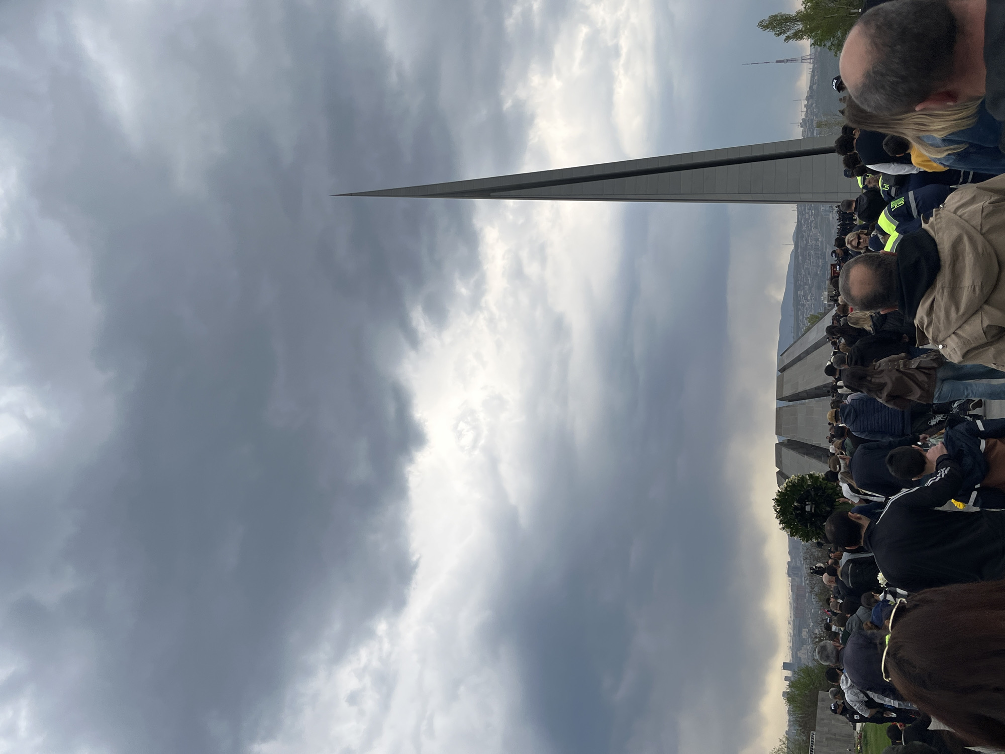 This image captures a solemn gathering of a large crowd at what appears to be a memorial complex under an overcast sky. The photograph is taken in a portrait orientation (if viewed upright), featuring a tall, slender, pointed obelisk-like monument that dominates the upper left part of the frame. A dense crowd of people, mostly adults seen from behind, occupies the right side of the image, extending into the foreground.
In the background, a cityscape unfolds, with a distinctive television tower visible on a distant hill, strongly suggesting the location is Yerevan, Armenia. Another architectural element, a wall-like structure composed of vertical gray concrete slabs, is visible on the far right, consistent with the design of the Tsitsernakaberd Armenian Genocide Memorial Complex.
The weather is cloudy, with patches of brighter sky indicating diffuse light, possibly late afternoon or a generally overcast day. The collective posture of the crowd and the setting imply an event of commemoration or remembrance, conveying a sense of solemnity. No discernible text is visible in the image.