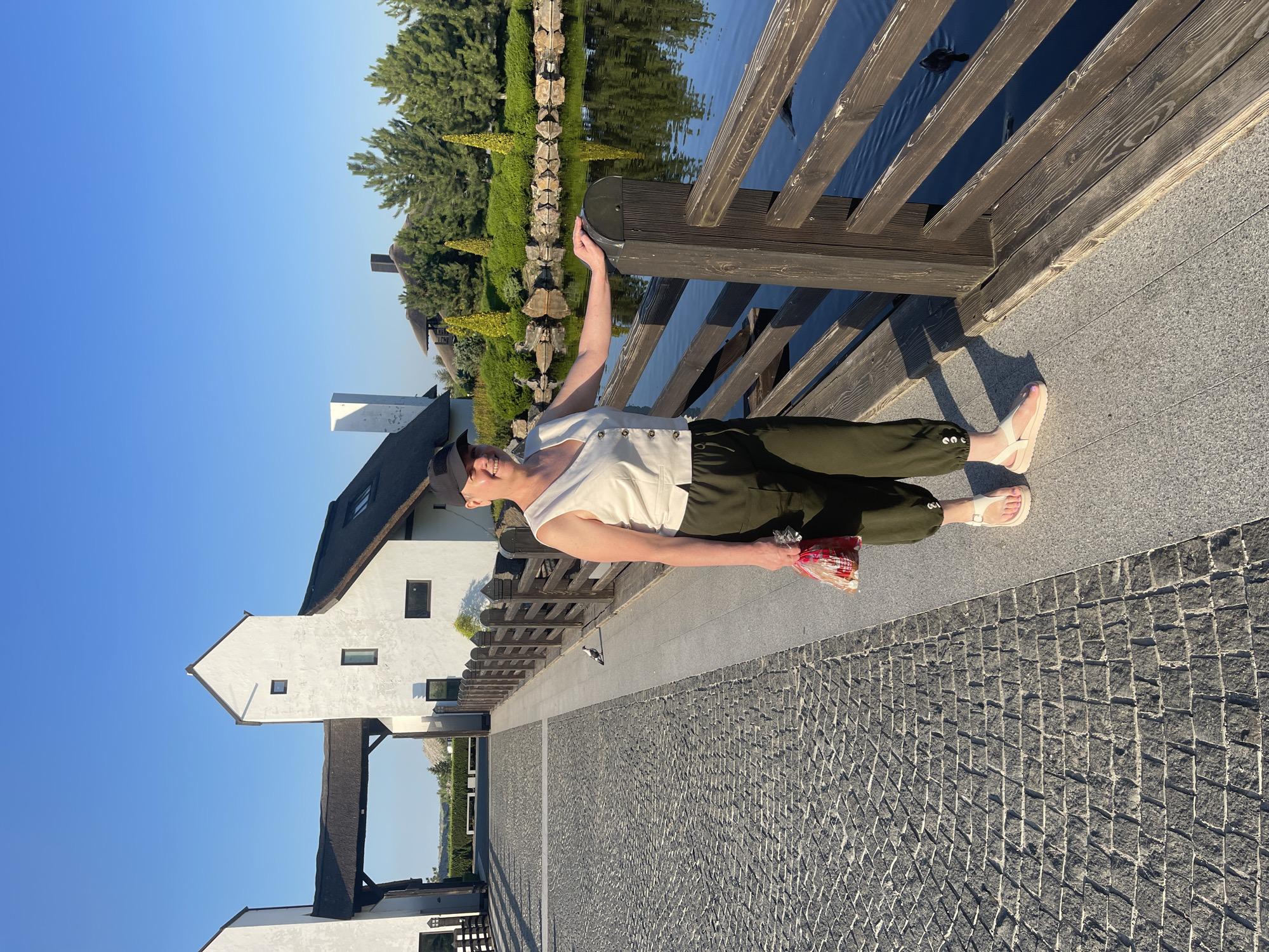 A woman wearing a white sleeveless vest, dark green pants, a black baseball cap, and white sandals stands on a paved pathway next to a wooden railing overlooking a body of water. She is holding a plastic bag filled with red items in her right hand. The pathway is made of dark cobblestones with a lighter gray border. In the background, there are white buildings with thatched roofs, a wooden fence, green trees, and lush landscaping around the water. The sky is clear and blue. A single bird is visible on the pathway to the left of the woman.
