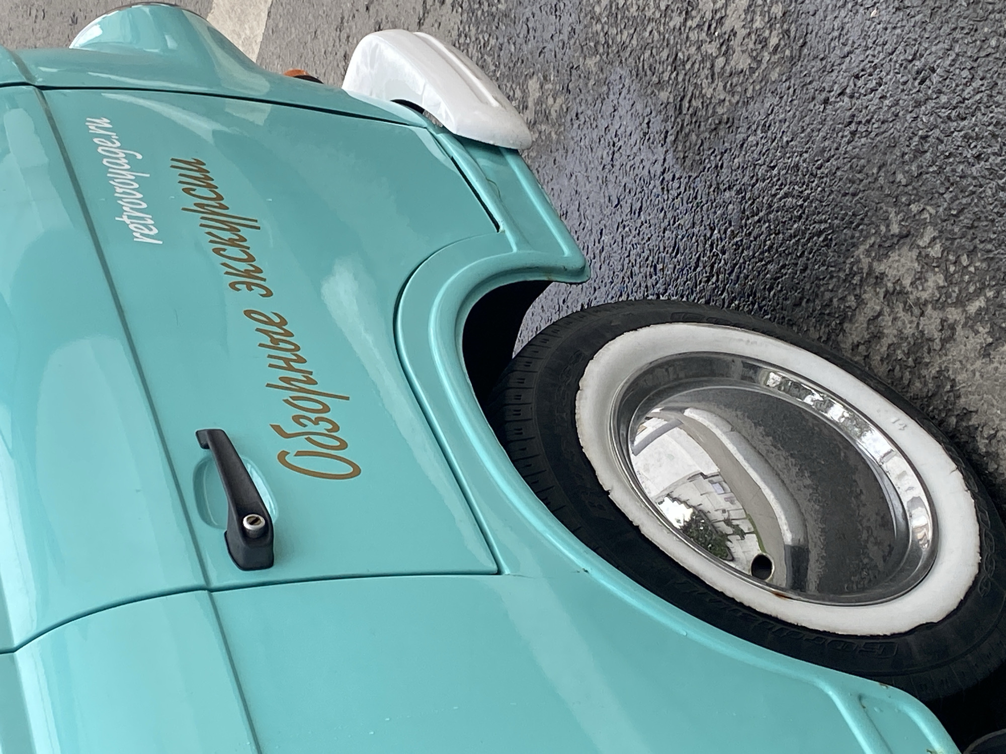 The image depicts a fragment of a light-blue vintage car, possibly located in the city of Шебунино, Russia, as suggested by the text "retrovoyage.ru" and "Обзорные экскурсии" (sightseeing tours) written on its side. The car features a white side mirror, a black door handle, and a tire with a white sidewall and chrome hubcap. The ground next to the car appears to be paved.
