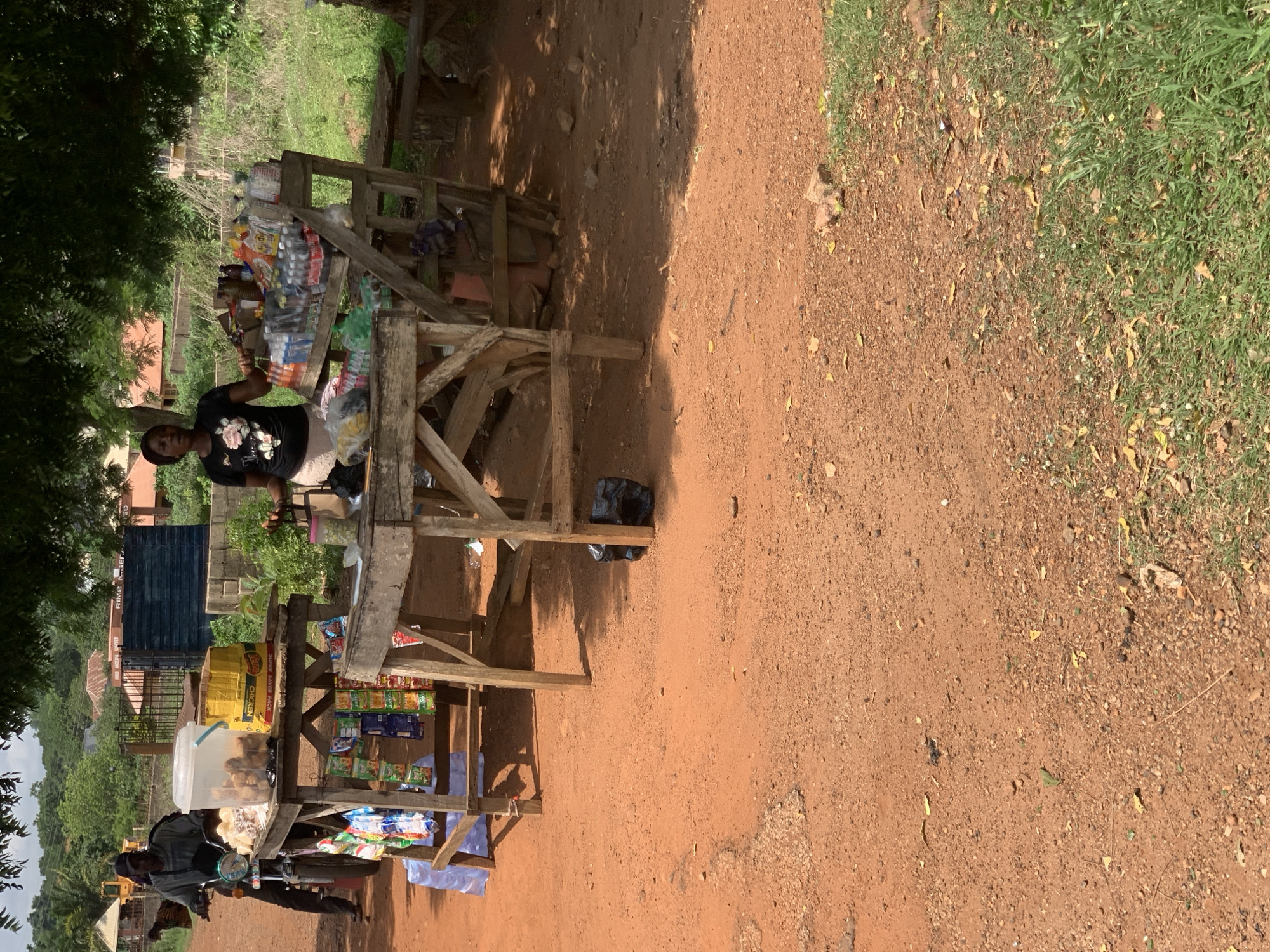 A street scene in Kabba, Nigeria shows a dirt road with a wooden roadside stand. The stand is stocked with packaged food items and beverages. A woman stands behind the stand. In the background, there are trees and a building. Another person sits on a parked motorcycle near the stand.