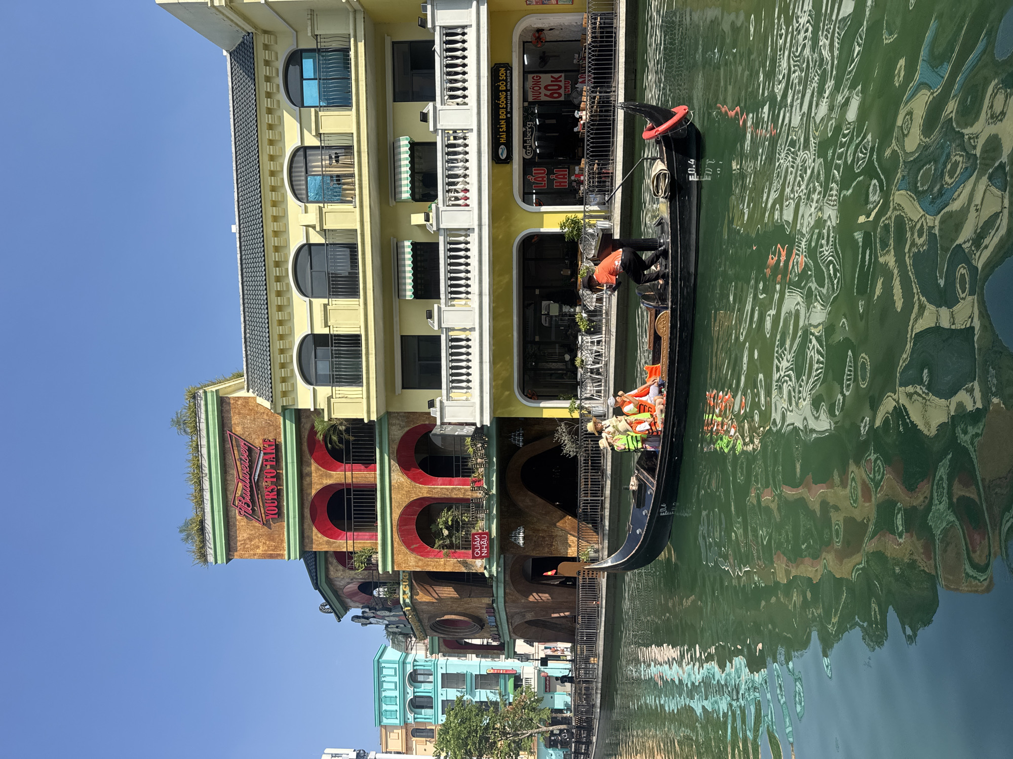 The image depicts a vibrant canal scene in Nghĩa Trụ Commune, Vietnam, featuring a black gondola gliding past brightly colored buildings under a clear blue sky. A gondolier, dressed in an orange shirt, stands at the stern, poling the boat. Several passengers, some wearing orange life vests, are seated in the gondola, which is marked "E0.4".
The multi-story buildings lining the canal exhibit varied architectural styles and colors, including yellow, reddish-brown, and light blue, with arched windows, balconies, and decorative railings. The green water of the canal reflects the buildings and the sunlight, creating shimmering patterns. Outdoor seating areas with tables and chairs are visible along the canal, suggesting restaurants or cafes.
The strong sunlight and clear sky indicate a bright day, likely midday or early afternoon. The architecture and the presence of a gondola evoke the atmosphere of Venice, but the visible text confirms the Vietnamese location.
Prominent signs on the buildings include:
* On a yellow building: "HẢI SẢN BƠI SỐNG ĐỒ SƠN" (Live Seafood Do Son), phone numbers "0968 636 868" and "0784 365 078", a "Carlsberg" logo, "LẨU HẢI" (Seafood Hotpot), and "NƯỚNG 60K" (Grill 60,000 VND).
* On a brown building: "Budweiser" and "YOURS TO TAKE".
* "QUÁN NHẬU" (Drinking/Eating establishment) is visible on another building with red arches.