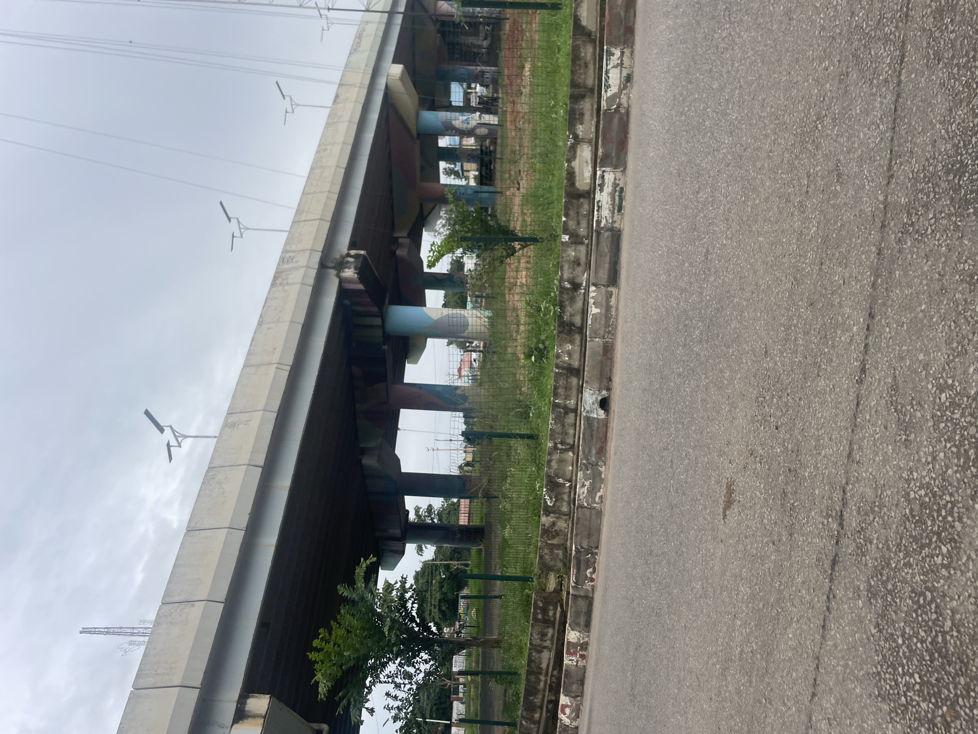 The image shows a section of an elevated highway in Jos, Nigeria. The highway is supported by large concrete pillars that are painted with colorful abstract designs. The area beneath the highway is overgrown with grass and has a green metal fence running along its perimeter. A road with a worn asphalt surface is in the foreground, with a concrete curb and drainage channel separating it from the fenced area. The sky is overcast, suggesting a cloudy day. Several streetlights are visible along the top of the highway. There are no people or vehicles clearly visible in the image.