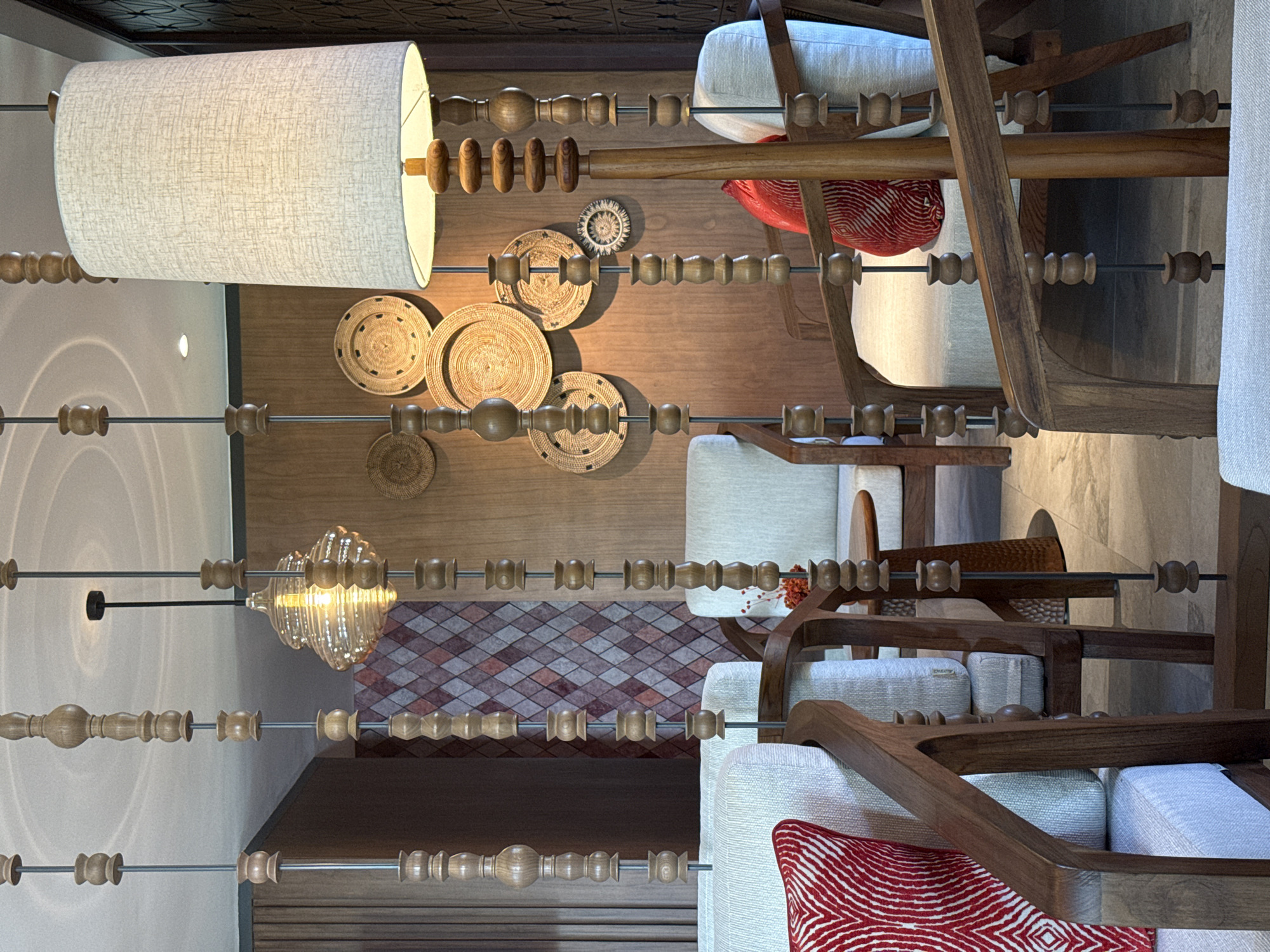 This image depicts a well-decorated indoor seating area, likely a lounge or lobby, situated in Kuta Selatan, Indonesia. The primary focus is on the furniture and decorative elements that create a warm and inviting atmosphere.
In the foreground, several wooden chairs with light-colored upholstery are visible. One chair prominently displays a red and white patterned cushion. The chairs are arranged around small, round wooden tables. A floor lamp with a textured beige lampshade stands to the right of the frame, its base adorned with wooden beads.
The walls are decorated with a variety of woven round plates and a patterned mosaic tile section, adding texture and visual interest to the space. Above, a decorative pendant light with a ribbed glass shade hangs, illuminated and casting a warm glow. Suspended from the ceiling are vertical wooden beaded strands, acting as decorative dividers or accents.
The flooring appears to be tiled in a neutral tone. The overall aesthetic suggests a blend of natural materials and artisanal craftsmanship, characteristic of upscale hospitality interiors in the region. There are no people visible in the image.
