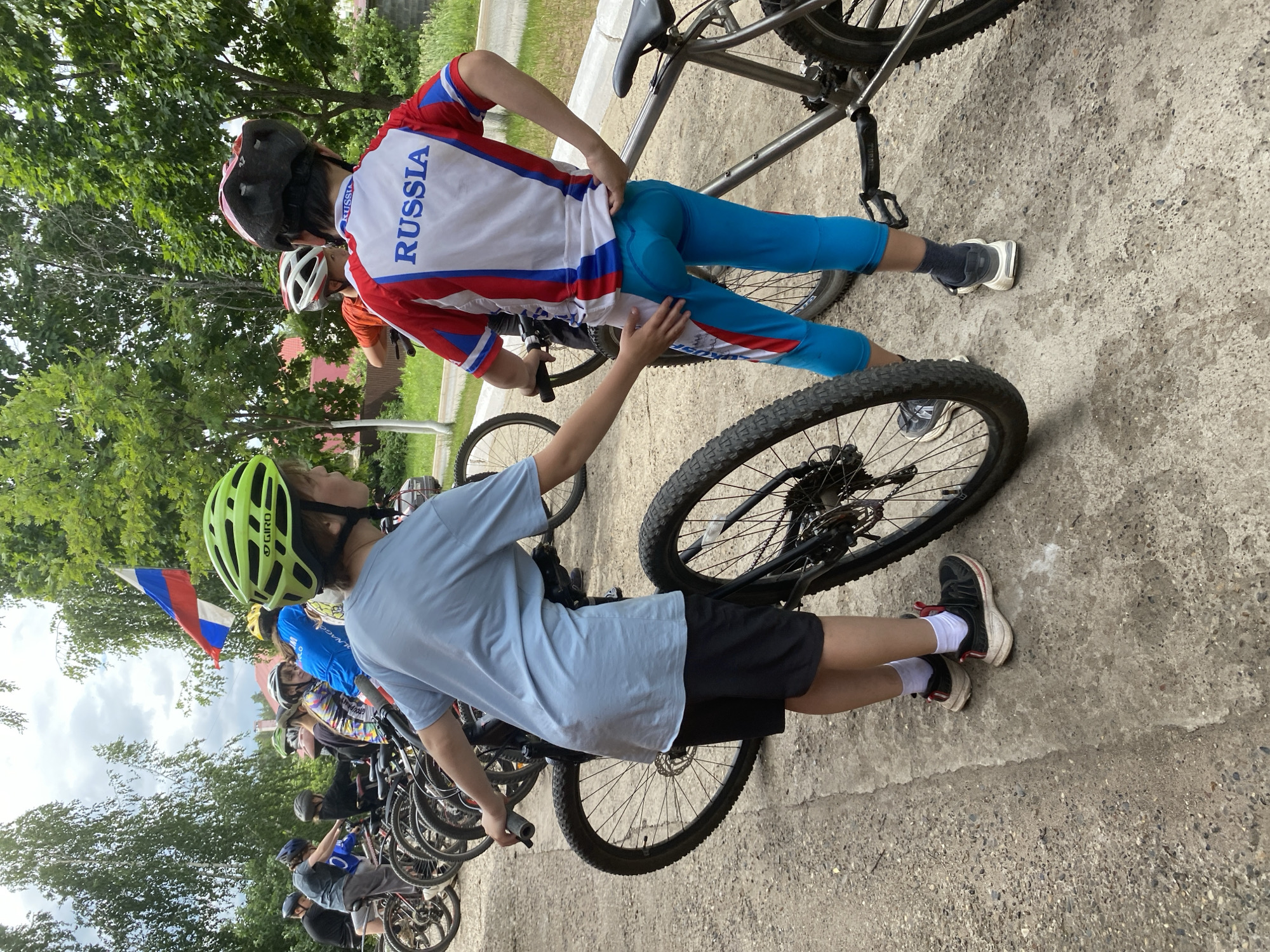 The image shows a group of children with bicycles, seemingly at an outdoor event. In the foreground, two boys are positioned. The boy on the left, wearing a light blue t-shirt and black shorts, is standing next to a bicycle, with his left hand on the handlebars and his right hand near the rear wheel. He is wearing a green helmet and black sneakers with white socks. The boy on the right, facing away from the camera, is wearing a red, white, and blue cycling jersey with "RUSSIA" printed on the back, and matching blue cycling shorts. He has a red and white helmet and dark sneakers. His right hand is touching the back of the boy in blue. Several other children and bicycles are visible in the background, lined up. A Russian flag is also visible, waving from a pole among the trees. The setting appears to be a gravel path or road with trees and buildings in the distance, suggesting a park or an open area within a town. The weather appears to be overcast. Based on the presence of the Russian flag and the word "RUSSIA" on the jersey, the location is likely in Russia. Without further information, pinpointing the exact location in Noviy, Russia, is not possible solely from the image content.