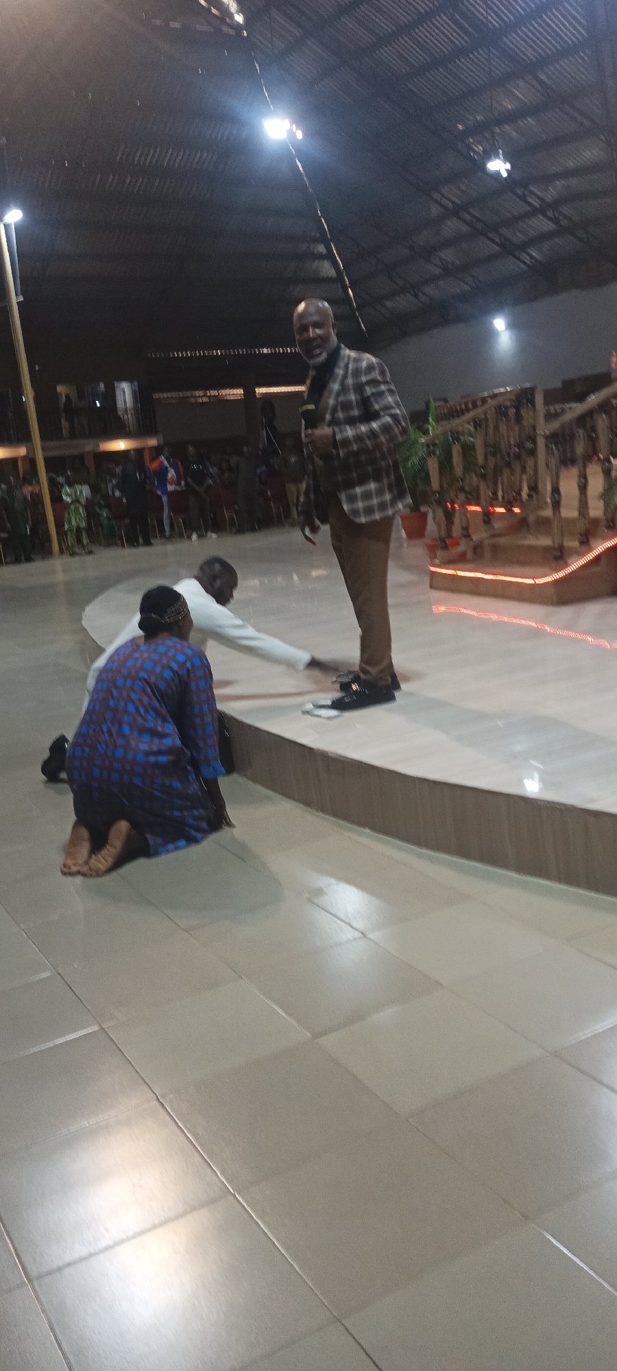 The image shows an indoor scene, likely a place of worship or an event hall, located on Rukuba Road, Nigeria. A man in a checkered blazer and brown trousers is speaking into a microphone, standing on a raised platform. In front of him, on the floor, two individuals are kneeling. One person, wearing a blue and brown patterned garment, is facing away from the camera, while another person in a white garment is reaching out towards the kneeling individuals. The environment appears to be a large open space with a corrugated metal roof and visible lighting fixtures. In the background, there are rows of chairs and an audience can be seen gathered. A staircase with decorative railings is also visible on the right side of the image. The floor is tiled with large square tiles.
