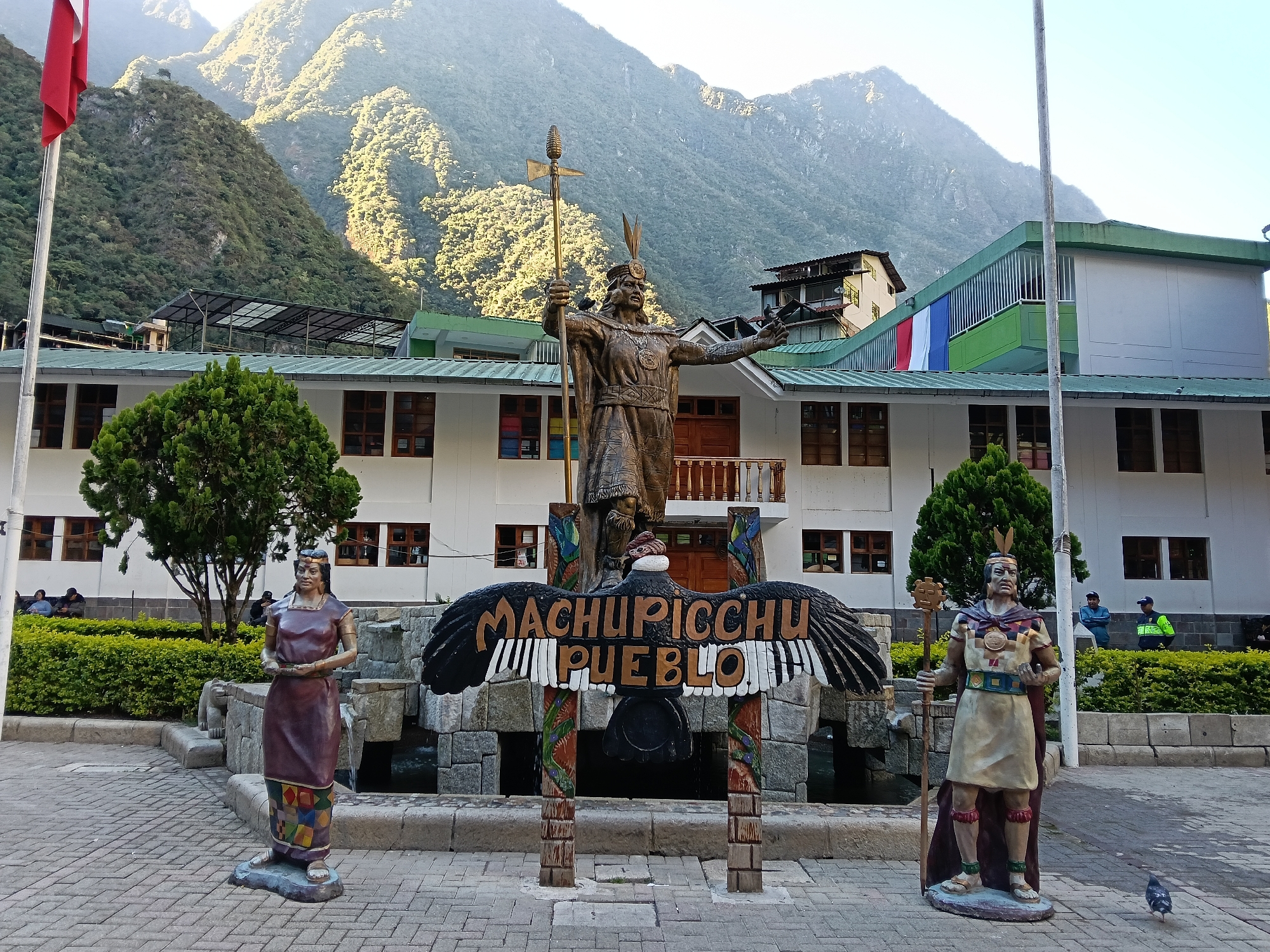 The image shows a plaza in the town of Machupicchu, Peru, located at the base of the Andes Mountains. In the foreground, from left to right, is a statue of a female figure, a sign reading "Machupicchu Pueblo" beneath a condor effigy, and a statue of a male figure. Behind these figures, there is a building painted white and green, with a Peruvian flag hung from a balcony. Behind the building are the mountains. The plaza floor is paved with light-colored bricks. There are people visible on a bench near a bush.