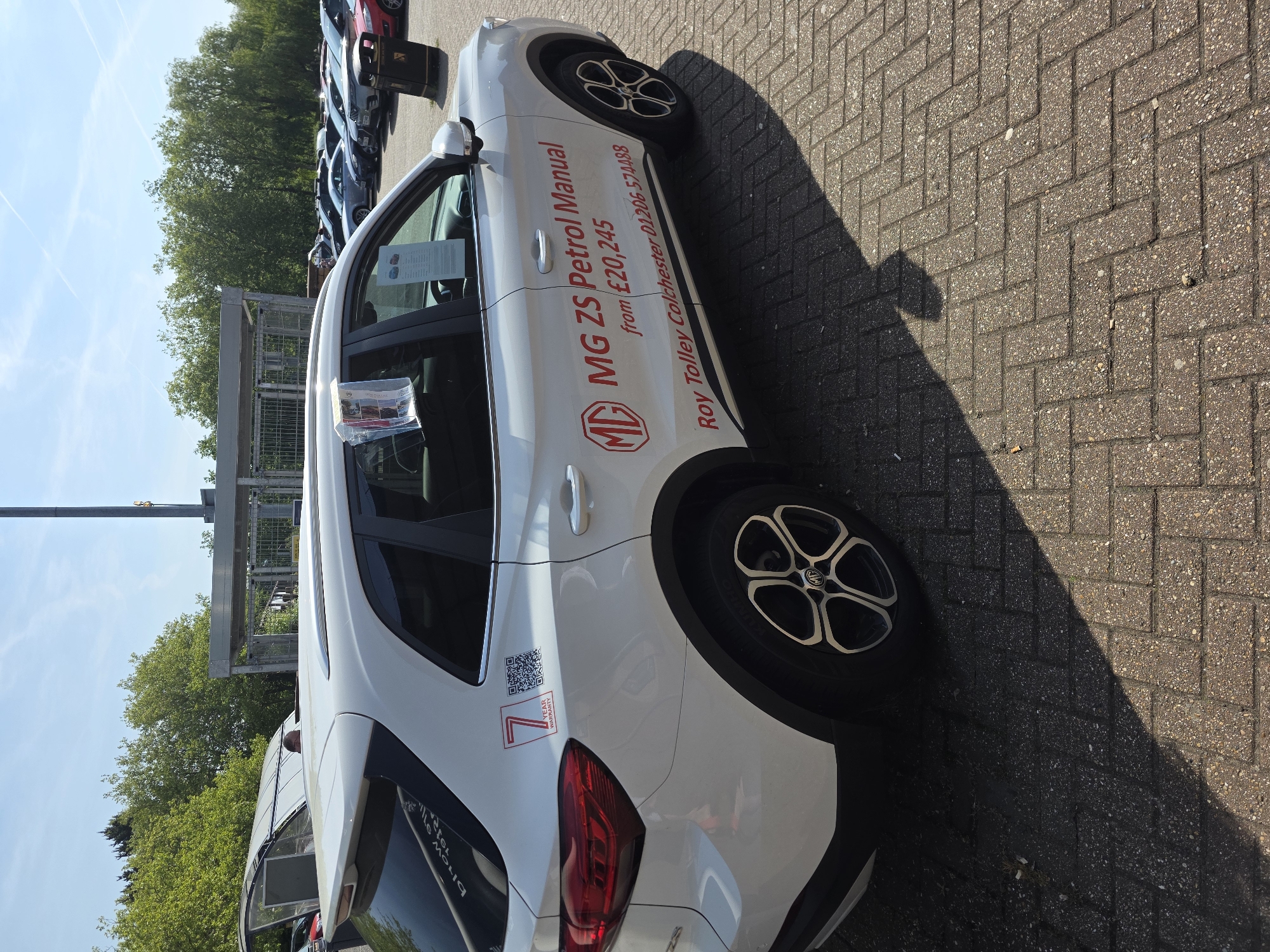 The image shows a white MG ZS Petrol Manual car parked on a paved surface, possibly at a car dealership. The car has "MG ZS Petrol Manual from £20,245" and "Roy Tolley Colchester 01206 574488" printed on its side, indicating that it is being sold at the Roy Tolley dealership in Colchester, United Kingdom. A "7 YEAR WARRANTY" sticker and QR code are visible near the car's rear light. Other cars are parked in the background, and trees are visible in the distance. The image is taken on a sunny day, as indicated by the shadow cast by the car.