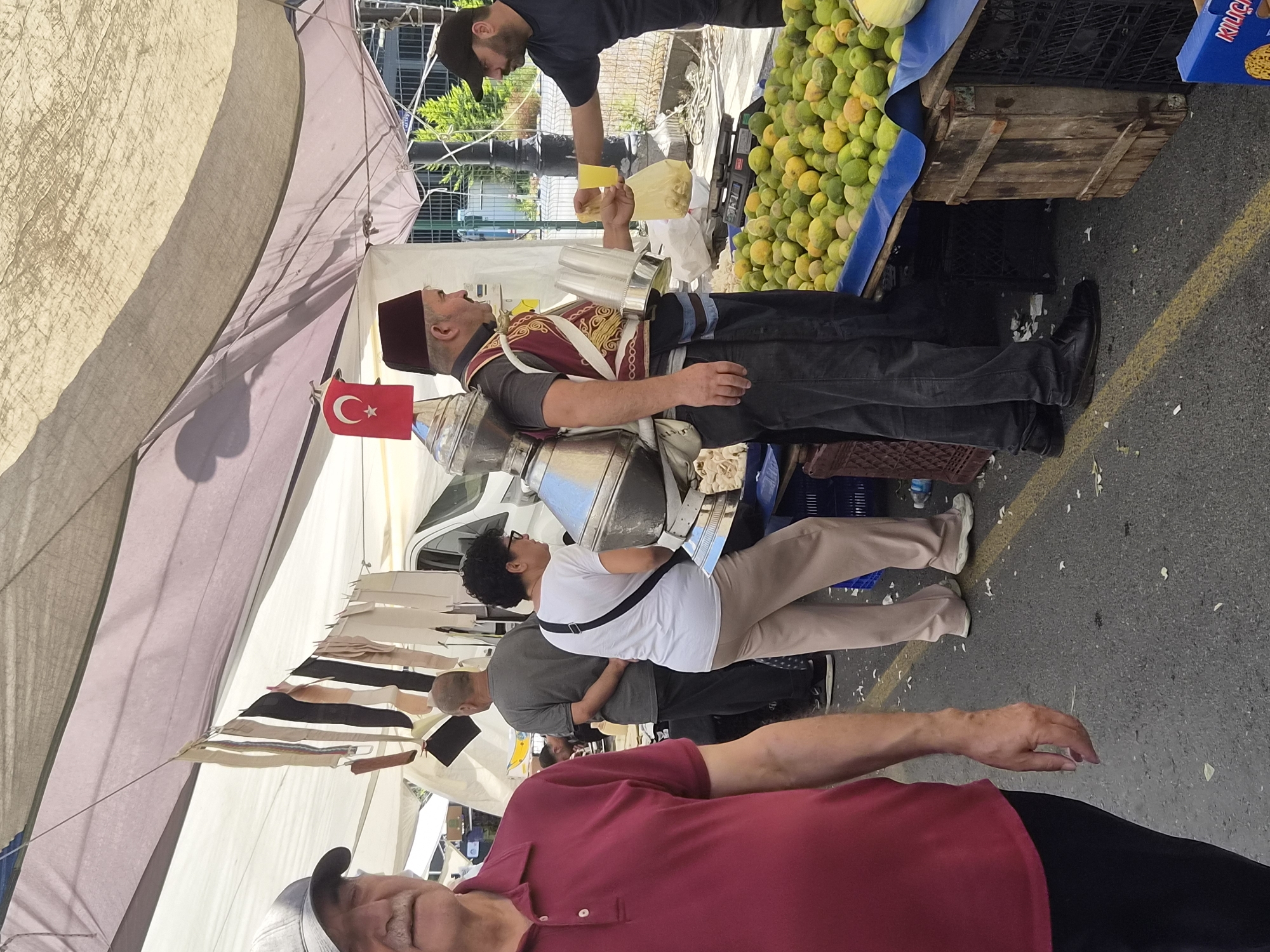 The image captures a street market scene, likely in Maltepe, Turkey, given the Turkish flag displayed. In the foreground, a man in a maroon polo shirt and black pants is partially visible, walking away from the camera. Behind him, a vendor wearing a traditional red fez and a decorative vest is serving a customer. The vendor carries a large, metallic, multi-tiered container on his back, characteristic of a traditional Ottoman-era street vendor, possibly selling sherbet or other beverages. He is handing a yellow cup to another man in a black t-shirt and cap, who is holding a plastic bag containing what appear to be lemons. In the background, a woman in a white t-shirt and beige pants stands near the vendor, looking towards the lemons. A large display of lemons is prominently featured in the mid-ground, piled high on a stall covered with a blue tarp. The market is set up outdoors, with a large fabric awning providing shade. Several people are browsing or working at stalls in the background, and a white van is visible behind some of the stalls. The ground is paved and littered with small white debris, possibly flower petals or paper.