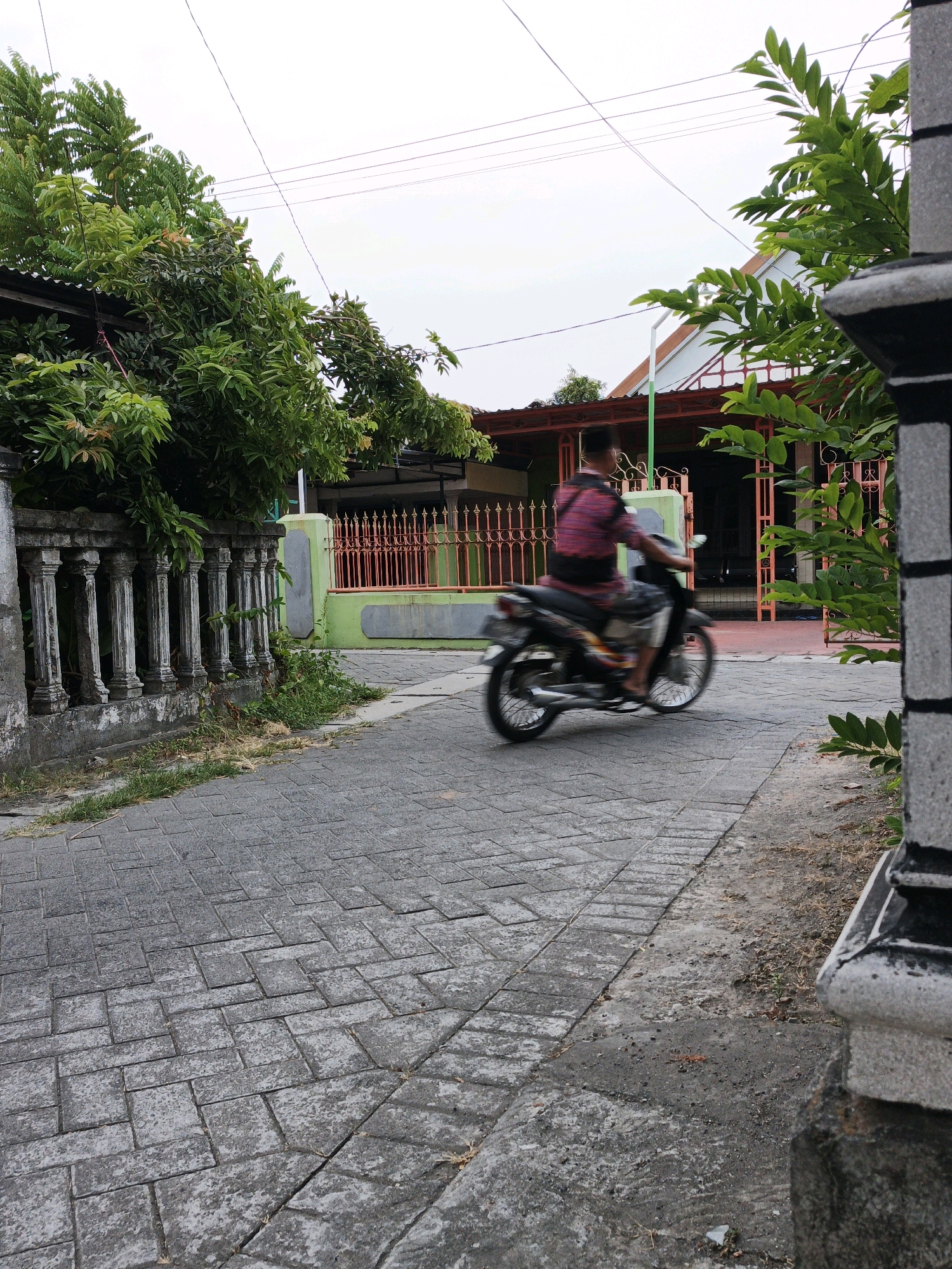 The image depicts a street scene in Ketegan, Indonesia. A person wearing a pink and black patterned shirt and dark pants is riding a motorcycle from left to right across the frame. The motorcycle and rider are blurred due to motion. The street is paved with gray interlocking concrete blocks. On the left side of the street, there is a stone fence with columns and green foliage growing from behind it. On the right side, part of a building's stone pillar is visible. In the background, there are houses with orange-tiled roofs and green and orange fences. The sky is overcast and pale. Power lines are visible stretched across the sky.