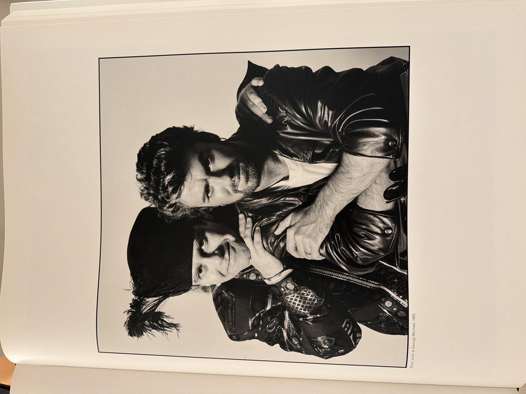 This is a black and white photograph of two men, Elton John and George Michael, posing together. They appear to be embracing or in a close embrace. Elton John is wearing a leather jacket and a white t-shirt, and George Michael is wearing a black hat with a tassel and a decorated jacket. The photograph is rotated 90 degrees counter-clockwise. The background is a plain, light color. Visible text at the bottom right of the photograph reads "Elton John & George Michael, 1985". The setting appears to be a studio or an indoor location with a neutral background. The mood seems playful and intimate. There are no visible cues for time of day or weather. The location context of Moscow, Russia, is provided externally and is not discernible from the image itself.