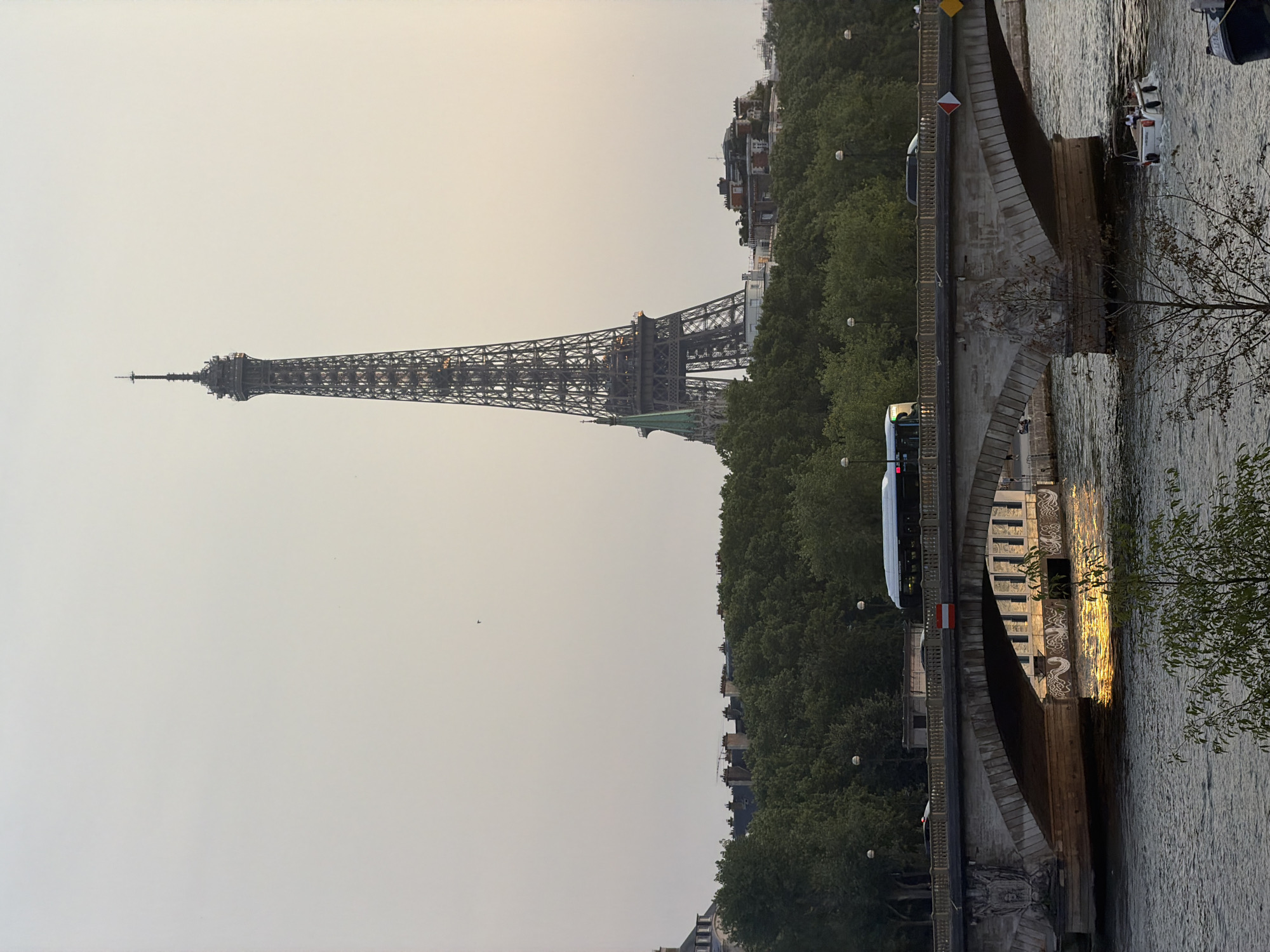 This image depicts a view of the Eiffel Tower in Paris, France, from across the Seine River. The Eiffel Tower, a tall iron lattice structure, dominates the background against a hazy, pale sky. In the midground, a stone bridge spans the river, with a bus and several cars visible on its roadway. The bridge has multiple arches, and its underside is illuminated by the reflection of the setting sun on the water. The Seine River flows in the foreground, with a small white boat with people on board moving across it. Lush green trees line the riverbank behind the bridge, and a cluster of Parisian buildings with distinctive mansard roofs can be seen peeking through the foliage in front of the Eiffel Tower. The scene is bathed in the warm, soft light of what appears to be either dawn or dusk.
