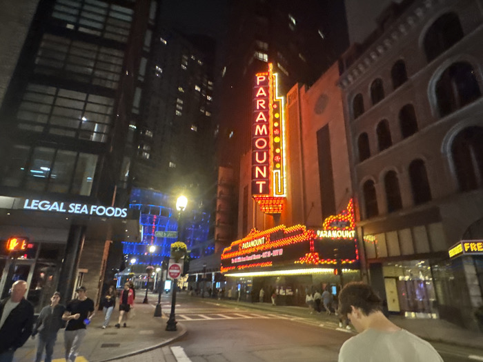 The image displays a nighttime urban scene on Washington Street in the Boston Theater District, Boston, Massachusetts. Prominently featured in the center-right is the Paramount Theatre, identifiable by its tall, vertical neon sign spelling "PARAMOUNT" in red letters with yellow border lighting, situated above an illuminated marquee also displaying "PARAMOUNT" with red and yellow lights. To the immediate left of the theater, a modern, multi-story glass-front building houses an illuminated "LEGAL SEA FOODS" sign at its base. Another dark building rises further left. To the right of the Paramount, an older, light-colored brick building with multiple arched windows is partially visible. The paved street occupies the lower portion of the image, with faint crosswalk markings. Several pedestrians, approximately 10-15 individuals, are visible on the sidewalks, walking in various directions. In the immediate foreground, a man with short dark hair, wearing a light-colored shirt, faces away from the camera, positioned slightly to the right. The scene is illuminated by streetlights, the glowing signage, and interior building lights, set against a dark night sky.