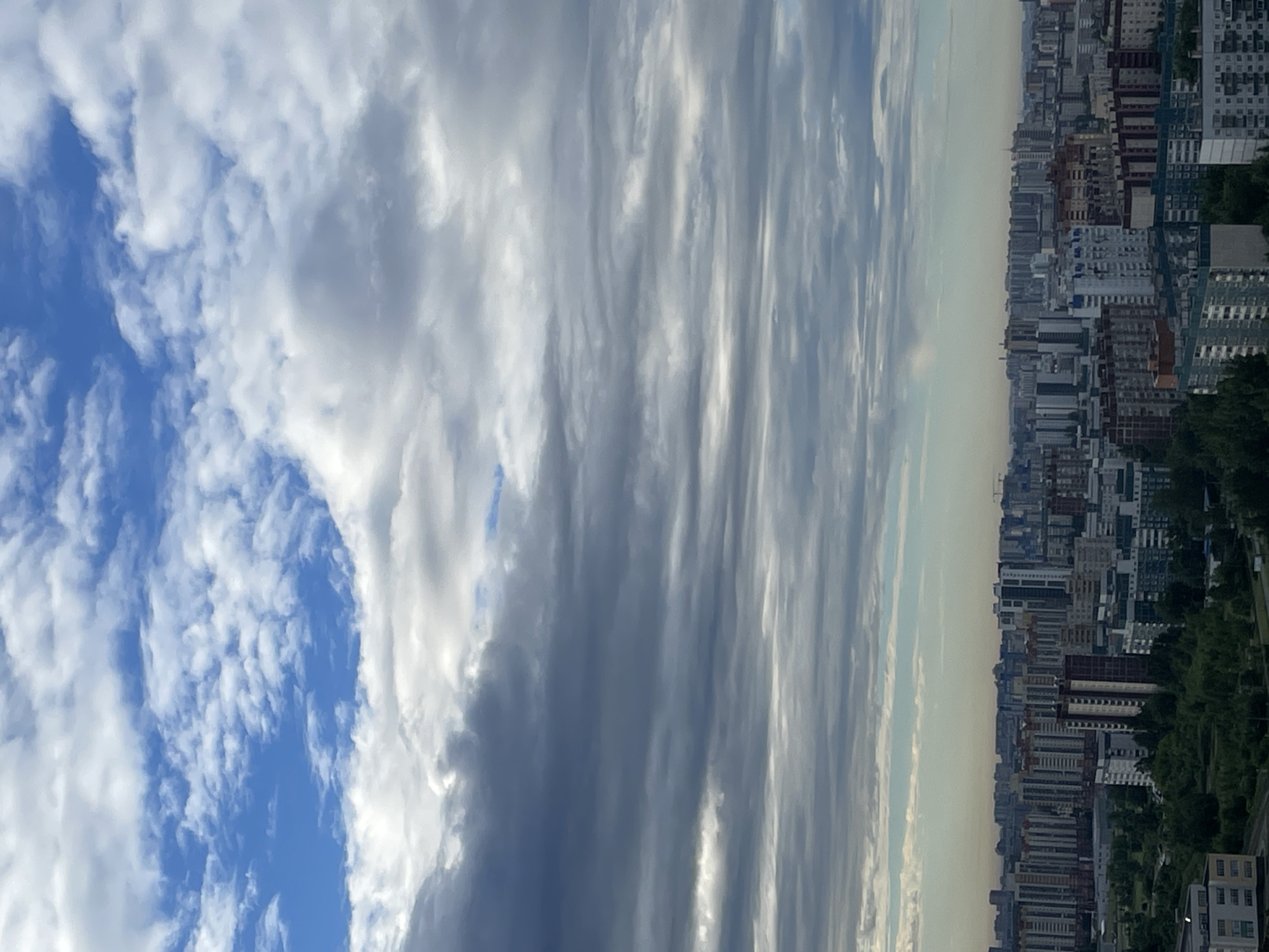 The image displays a cityscape viewed from above, with a significant portion dedicated to the sky and clouds. The scene is oriented vertically, making it appear as if the viewer is looking sideways at the horizon. On the right side of the frame, a dense urban environment unfolds, characterized by numerous multi-story buildings of varying architectural styles and colors, packed closely together. The buildings are a mix of residential and possibly commercial structures, with many featuring prominent windows and balconies. Hints of greenery, such as trees and park areas, can be seen interspersed among the buildings, suggesting a city with some urban planning for green spaces.
The sky occupies the left and central portions of the image and is filled with a dramatic display of clouds. These clouds range from bright white, suggesting sunlight reflecting off them, to darker, more ominous gray tones, indicating depth and density. The cloud formations appear layered and streaky, with some areas showing wispy, cirrus-like patterns and others presenting more robust, cumulus formations. The contrast between the vast sky and the man-made city creates a striking visual.
The time of day appears to be either early morning or late afternoon, as the light quality suggests the sun is low on the horizon, casting a diffused light. The weather seems overcast or partly cloudy, with the dramatic cloud cover dominating the sky. There are no visible people or specific activities taking place, and no text is present in the image. The overall impression is one of a sprawling urban landscape under a dynamic sky, possibly captured during a transition in weather conditions or at a time of day with unique lighting.