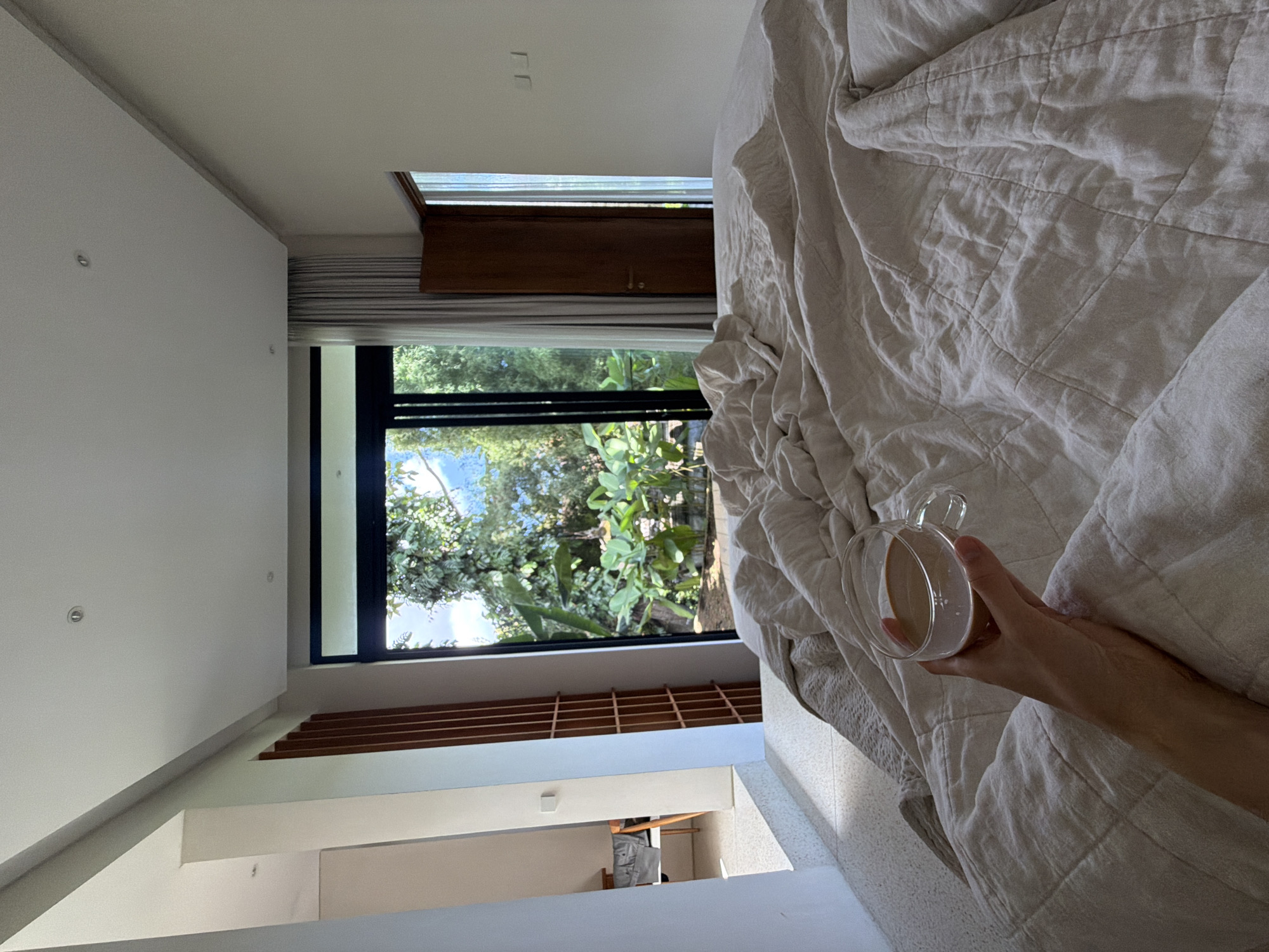 A vertical indoor shot shows a person's arm holding a clear glass mug with a light brown liquid inside, resting on a wrinkled, light gray quilt on a bed. The bed is near a large, floor-to-ceiling black-framed window showing lush green foliage and trees under a blue sky. Part of the window has a horizontal, dark wooden panel above it. The interior walls are white, with modern architectural details. A glimpse into another room reveals a person and furniture. The location is likely in a home in Mas, Indonesia, as specified.