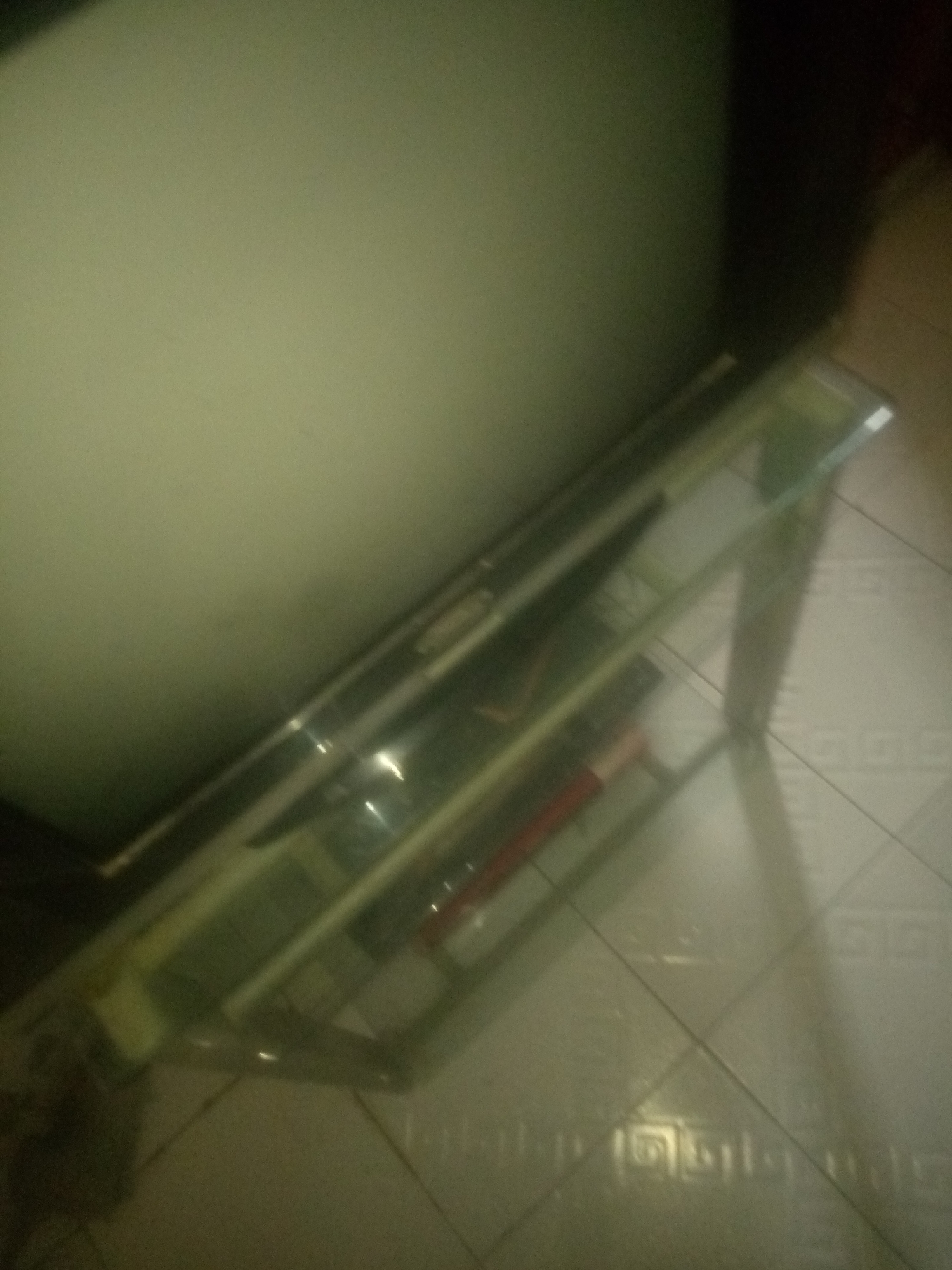 This is a blurry, close-up, indoor shot of a glass shelf unit, possibly part of a TV stand or entertainment center. The shelf is made of clear glass and supported by what appears to be a metal frame, though the frame is not clearly visible. The shelf is angled diagonally across the frame, from the lower left to the upper right.
On the shelf, there are several objects, but they are all indistinct due to the blurriness of the image. It is possible to discern some shapes, including what looks like a red cylindrical object, perhaps a pen or a marker, near the front of the shelf. There are also longer, more elongated objects that could be electronic devices or other items stored on the shelf.
The background consists of a light-colored, possibly cream or beige, wall on the left side of the frame. The floor is tiled with square tiles that have a subtle pattern. The lighting is dim, creating shadows and reflections on the glass shelf, further obscuring the details of the objects. The overall impression is of a dimly lit room, with the focus on the glass shelving unit. There is no visible text in the image. The time of day and weather conditions are not discernible.