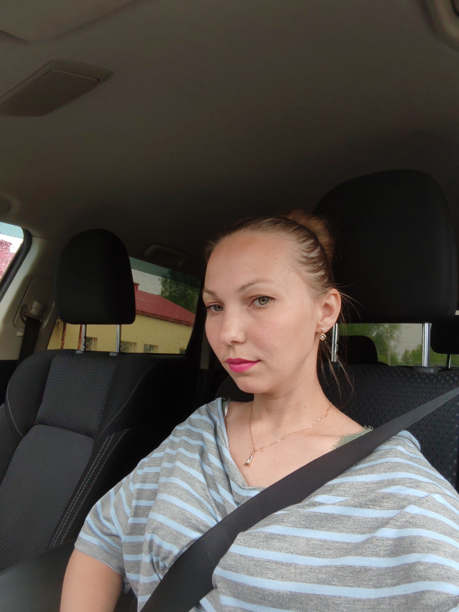 The image captures a woman wearing a striped gray and light blue top, seated in the driver's seat of a car with a seatbelt fastened across her chest. She has her hair pulled back in a bun, wears earrings and a gold necklace, and has pink lipstick on. The car's interior features black seats and headrests. Outside the car window, rain is visible, and a building with a red roof and yellow walls can be seen. The location is identified as Чухлэм, Russia.