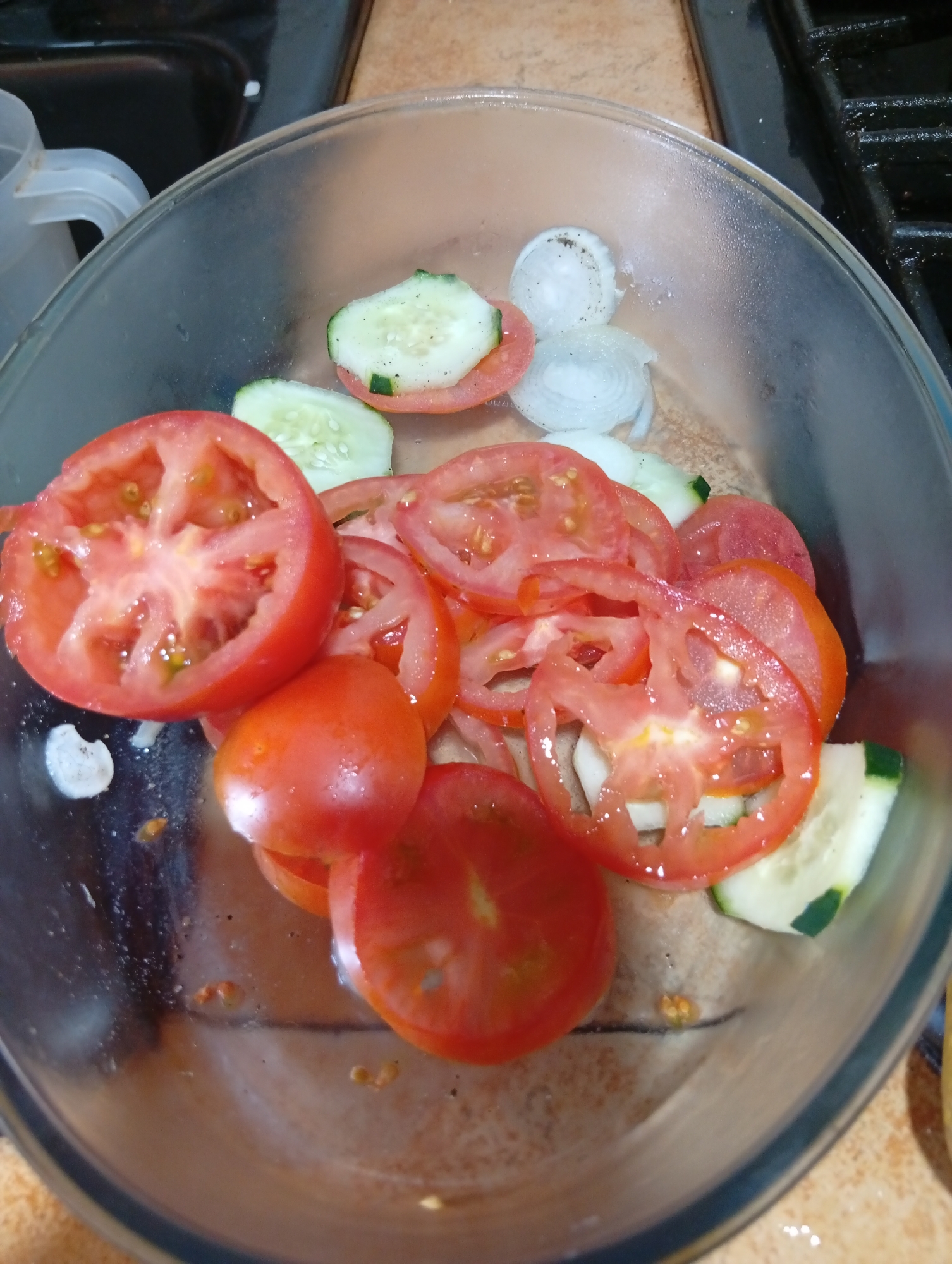 A clear glass bowl contains a salad made of sliced tomatoes, cucumbers, and onions. The ingredients are layered and mixed within the bowl, with some appearing to be more freshly cut than others. The salad appears to be on a surface that is a mix of tile and possibly a countertop. The background shows a stovetop and a plastic container with a handle, suggesting a kitchen environment. The overall scene suggests a preparation or consumption of a simple salad. The location is identified as San Antonio de Los Altos, Venezuela.